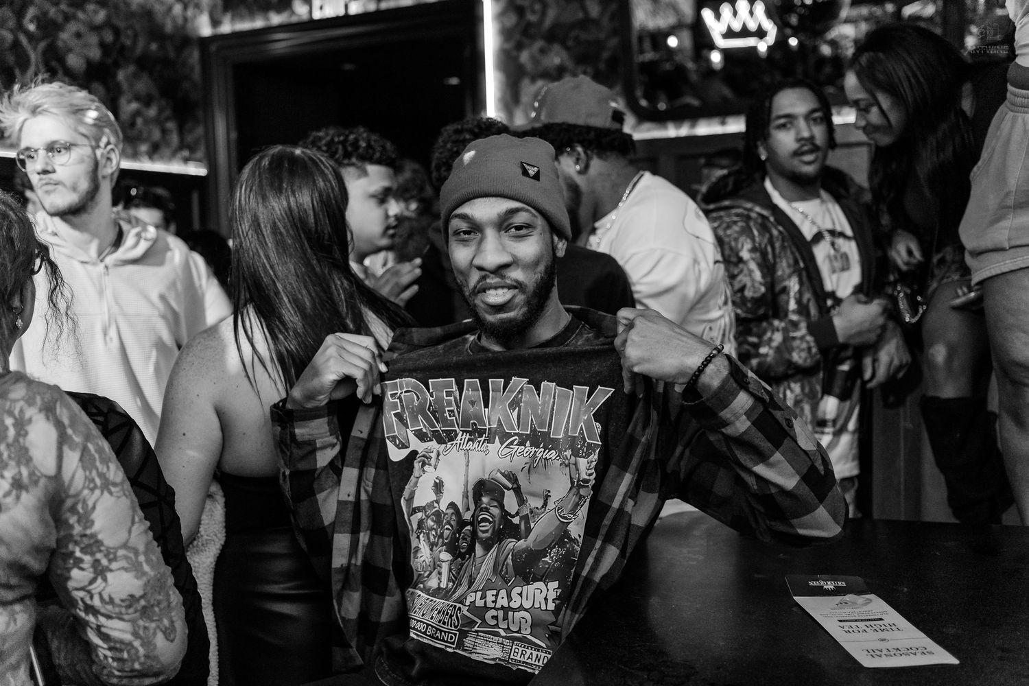 Man in beanie proudly displaying graphic tee at crowded bar, surrounded by tattooed guests in black and white photo.