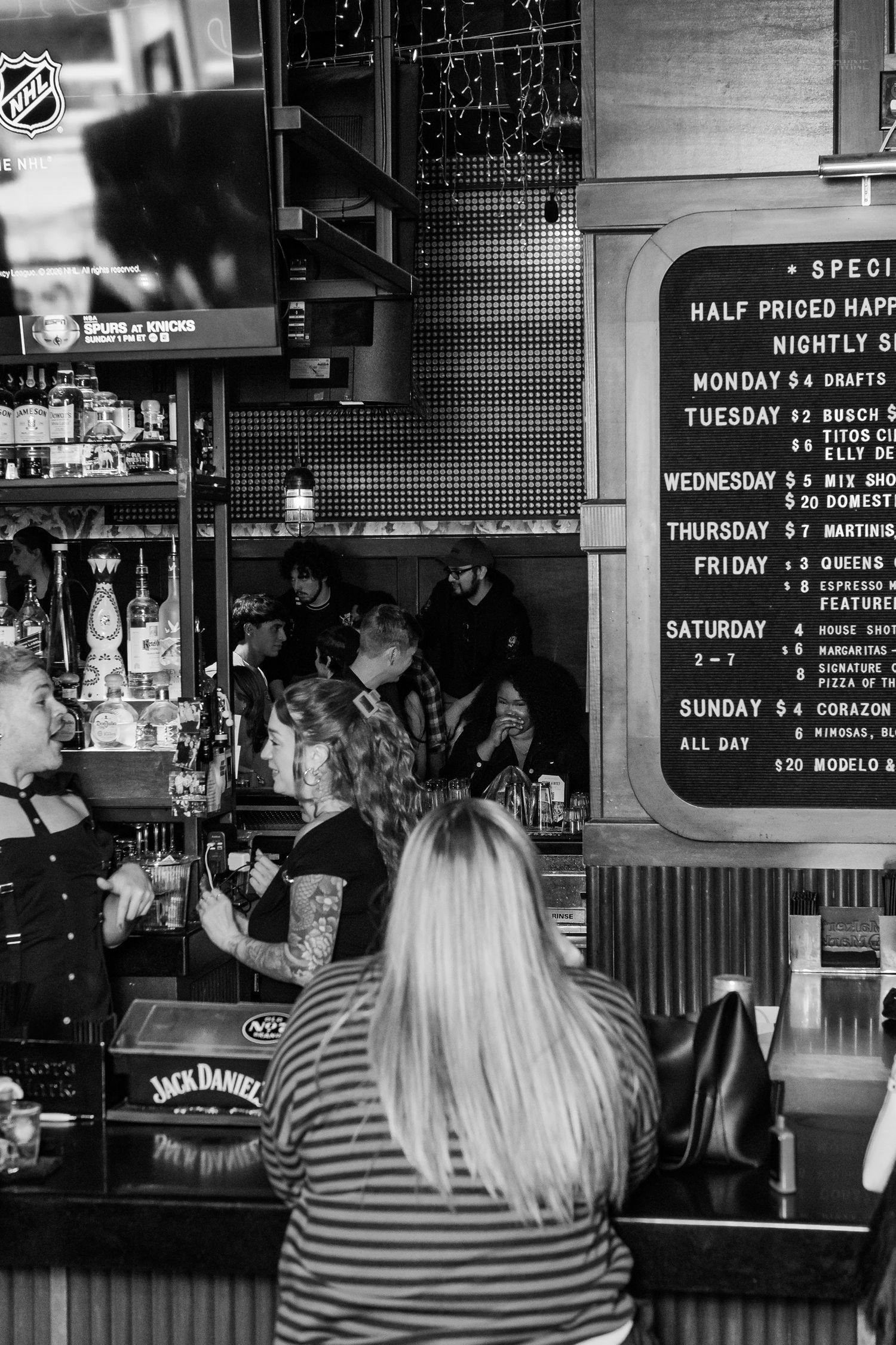 Patrons gather at a bar featuring Jack Daniels branding and a happy hour specials chalkboard menu.