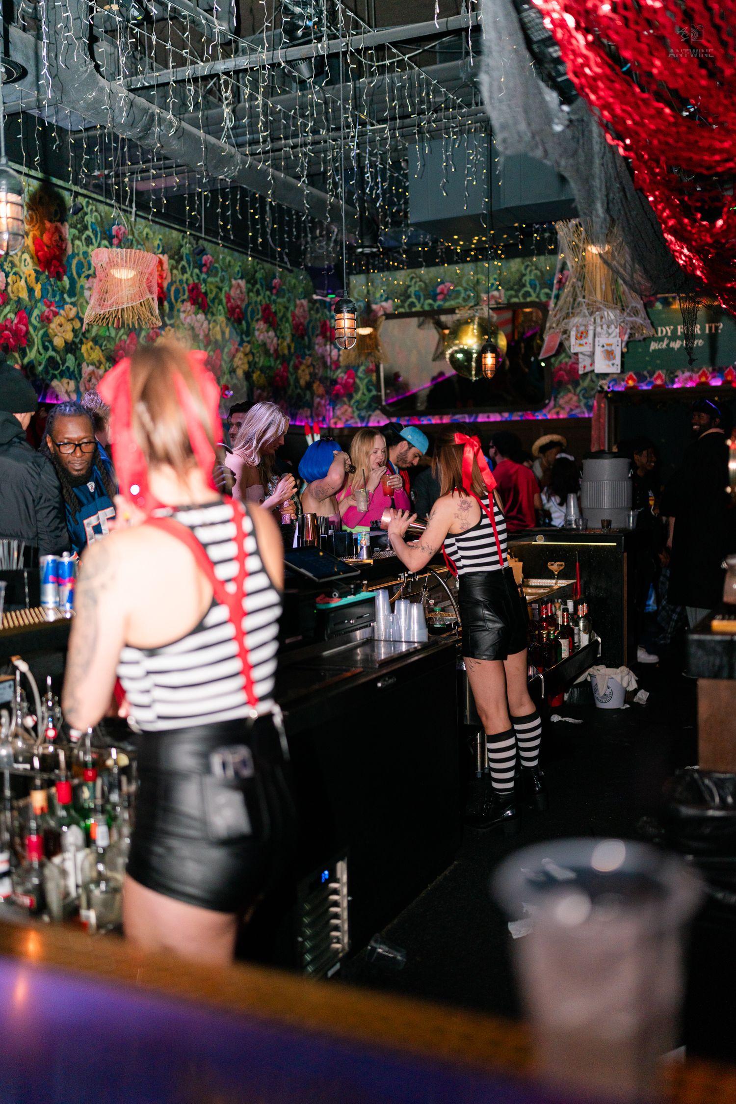 Bartenders in costumes serving drinks at a vibrant nightclub bar decorated with colorful floral decor.