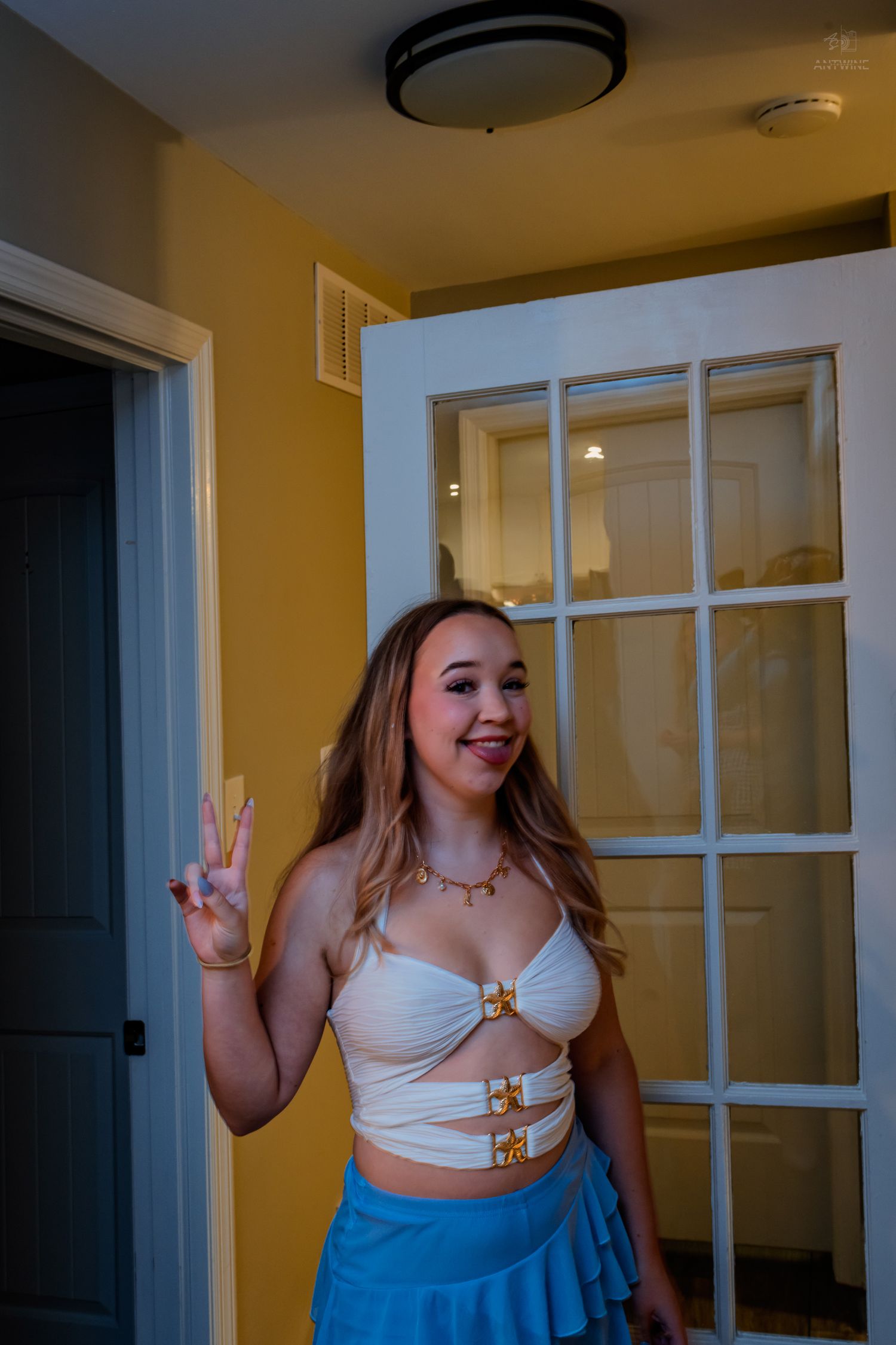 Young woman in white crop top and blue skirt making peace sign inside yellow-walled home.