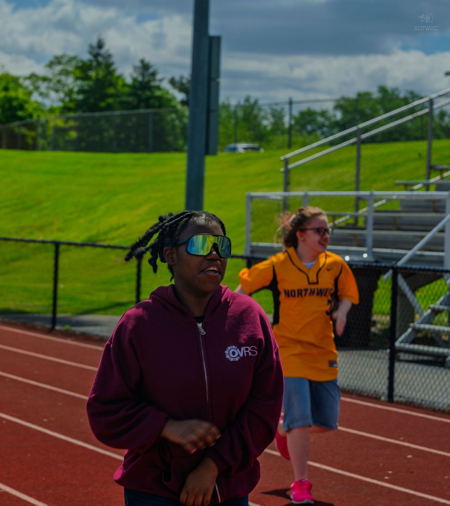 Two athletes on a red track, one in a maroon hoodie with sunglasses, another in a yellow jersey, with bleachers behind.