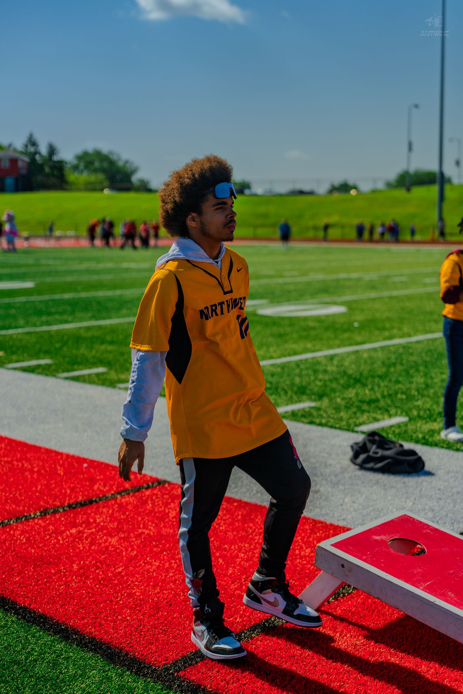 A young man in a yellow jersey plays cornhole on a red turf field at an outdoor sports event.