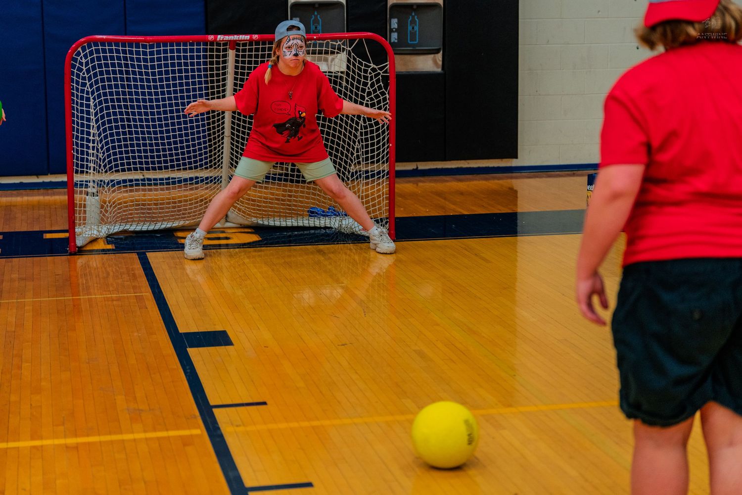 Children playing indoor soccer in a gymnasium, with a girl in red guarding a small hockey goal on a hardwood floor.