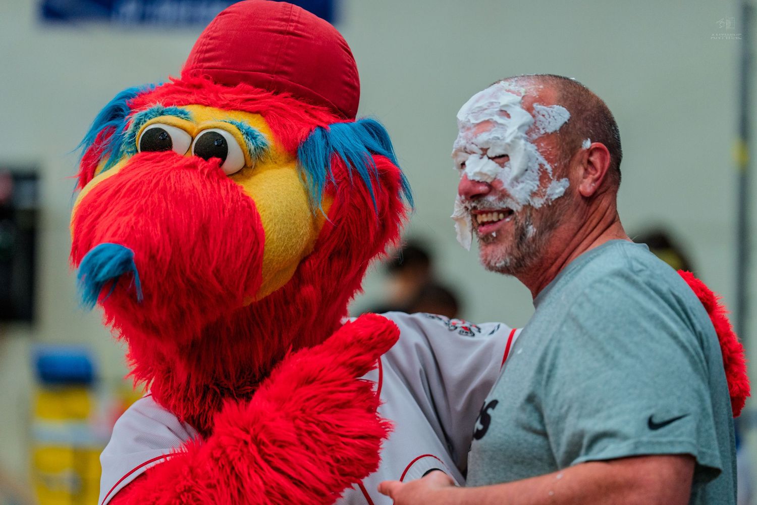 A man with whipped cream on his face laughs with the Phillie Phanatic mascot at a fun baseball event.