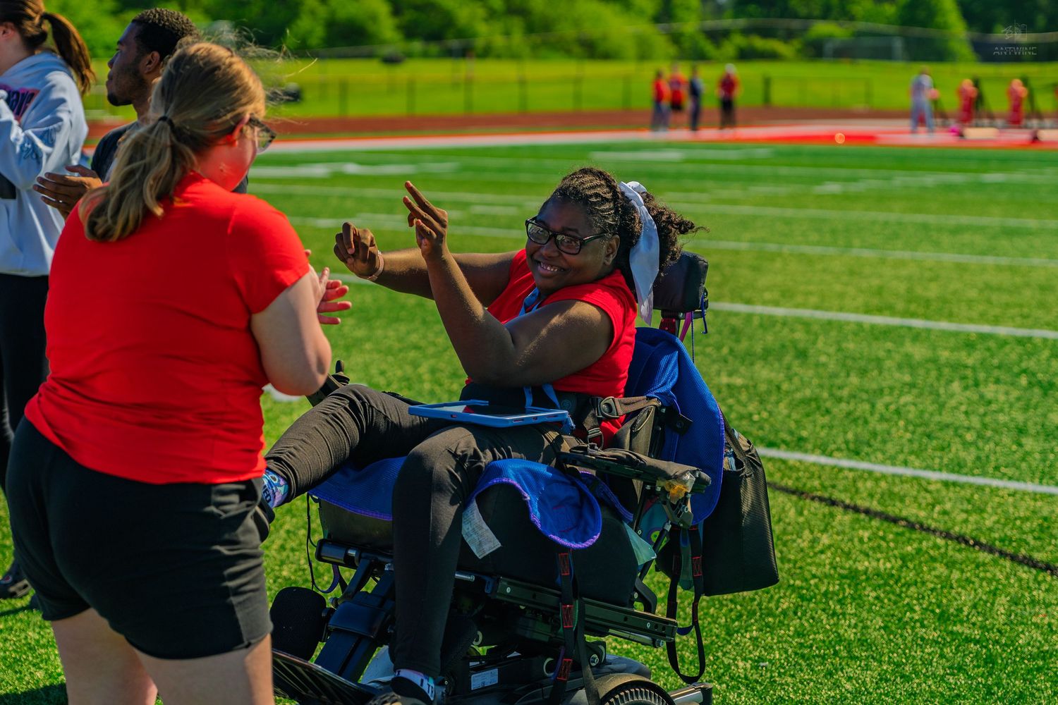 A young woman in a wheelchair high-fiving a person in a red shirt on a green turf athletic field.