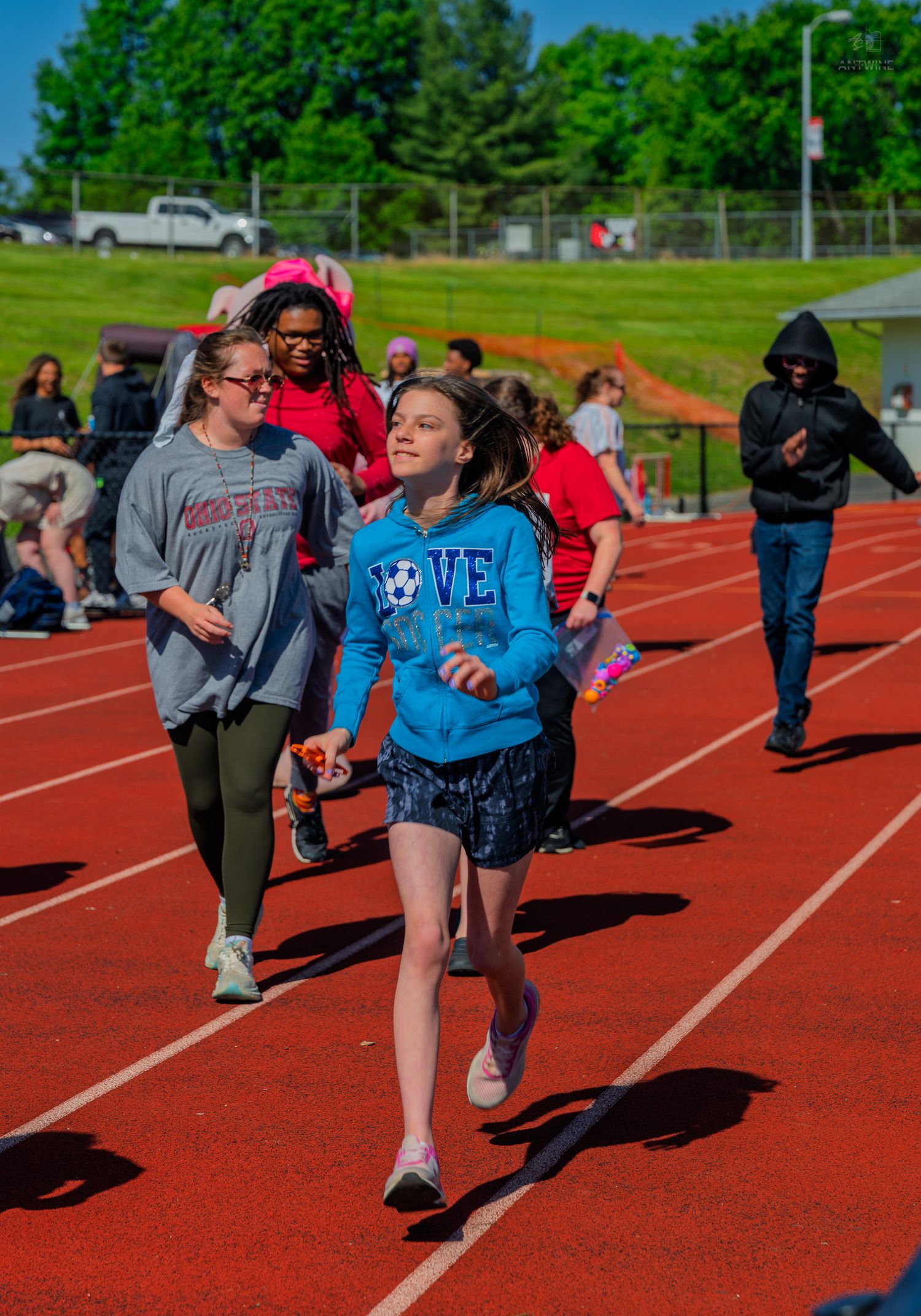 Young girl in blue LOVE shirt running on red track at school field day event with other students.