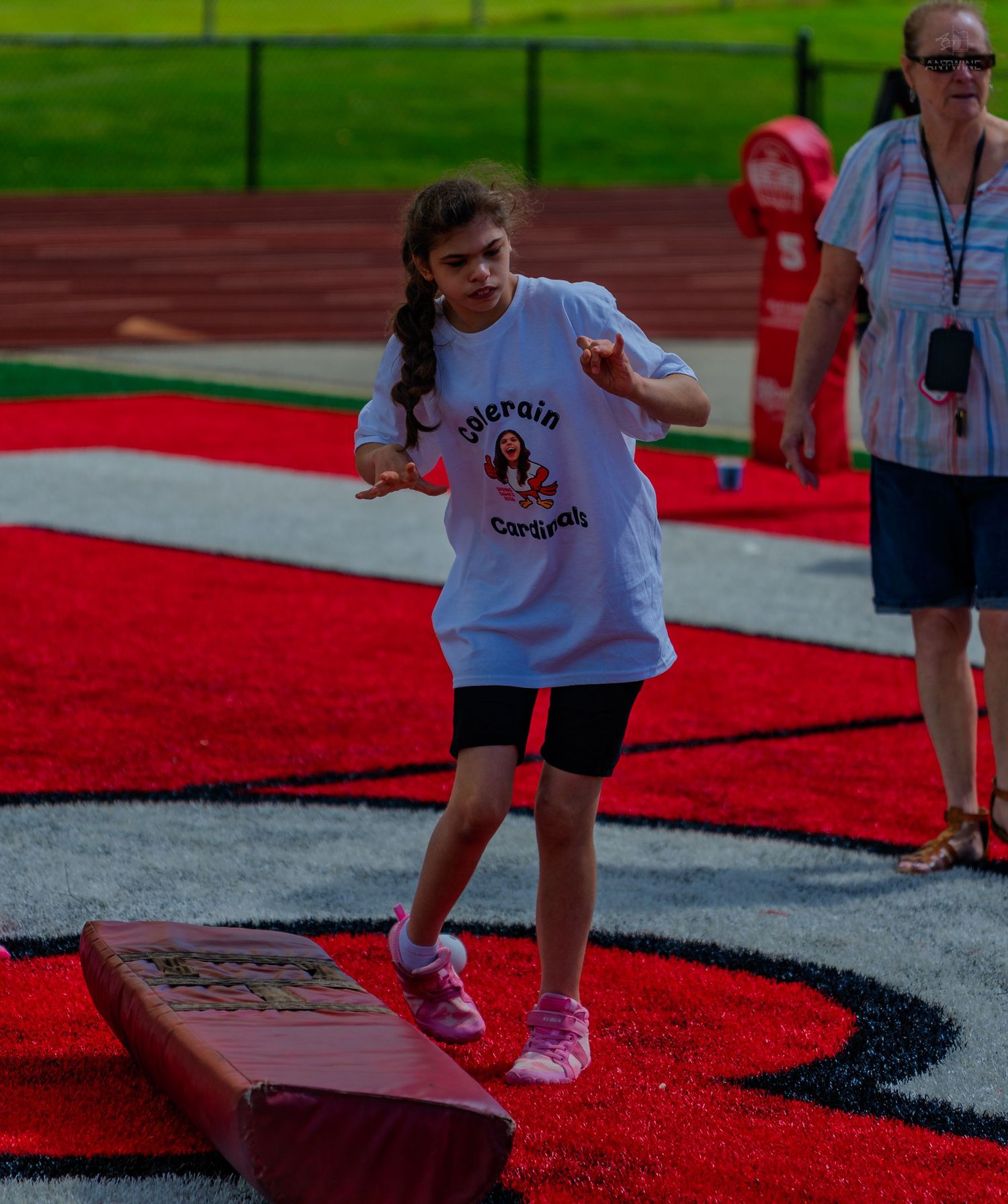 Young girl balancing on a beam outdoors at a red track field during a school activity event.