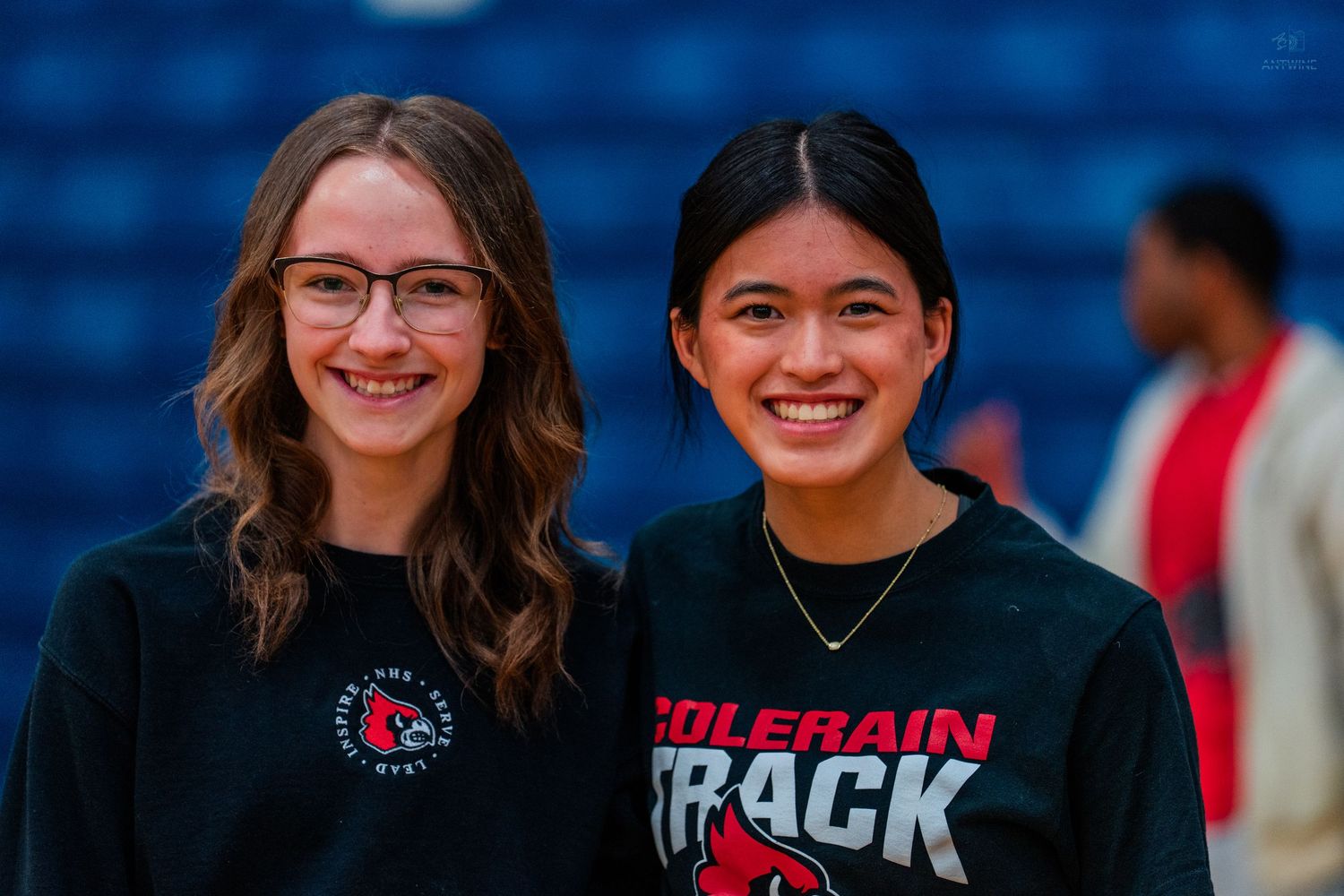 Two smiling teenage girls wearing Colerain Track black shirts standing together in a gymnasium.