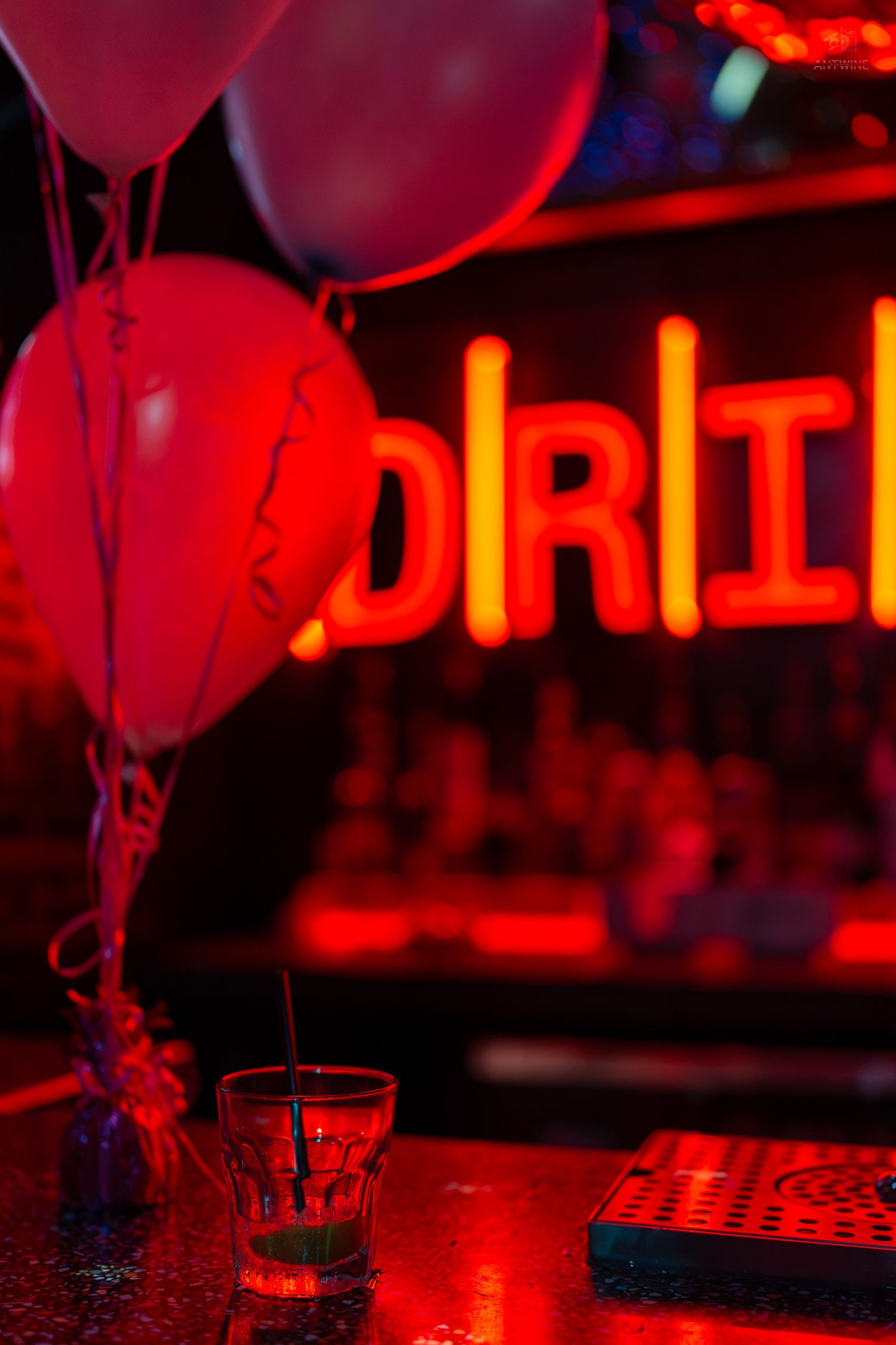 Red balloons and a shot glass on a bar counter with glowing neon 'DRINK' sign in the background.