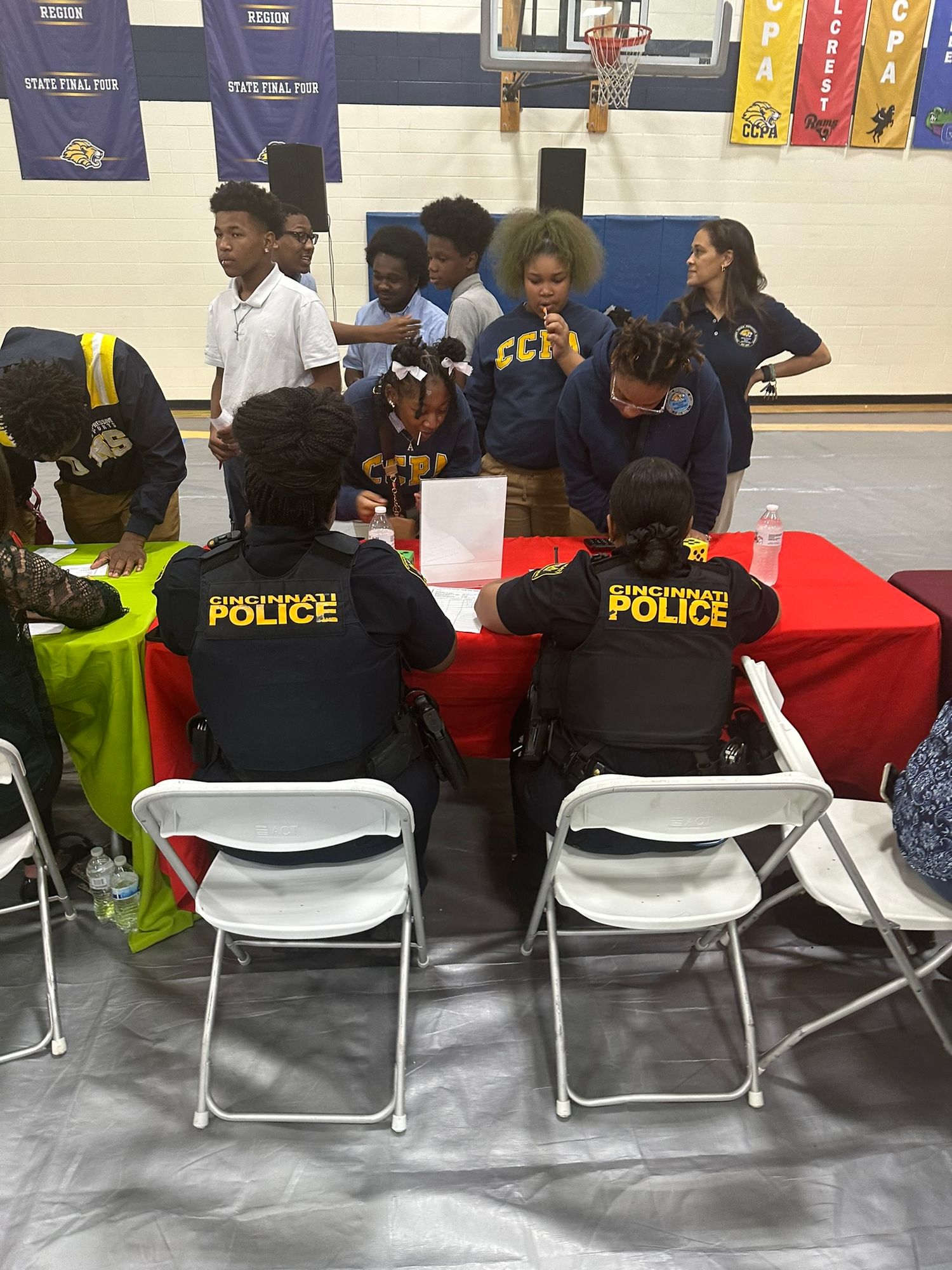 Cincinnati Police officers sit at a table during a school career fair event in a gymnasium with students.