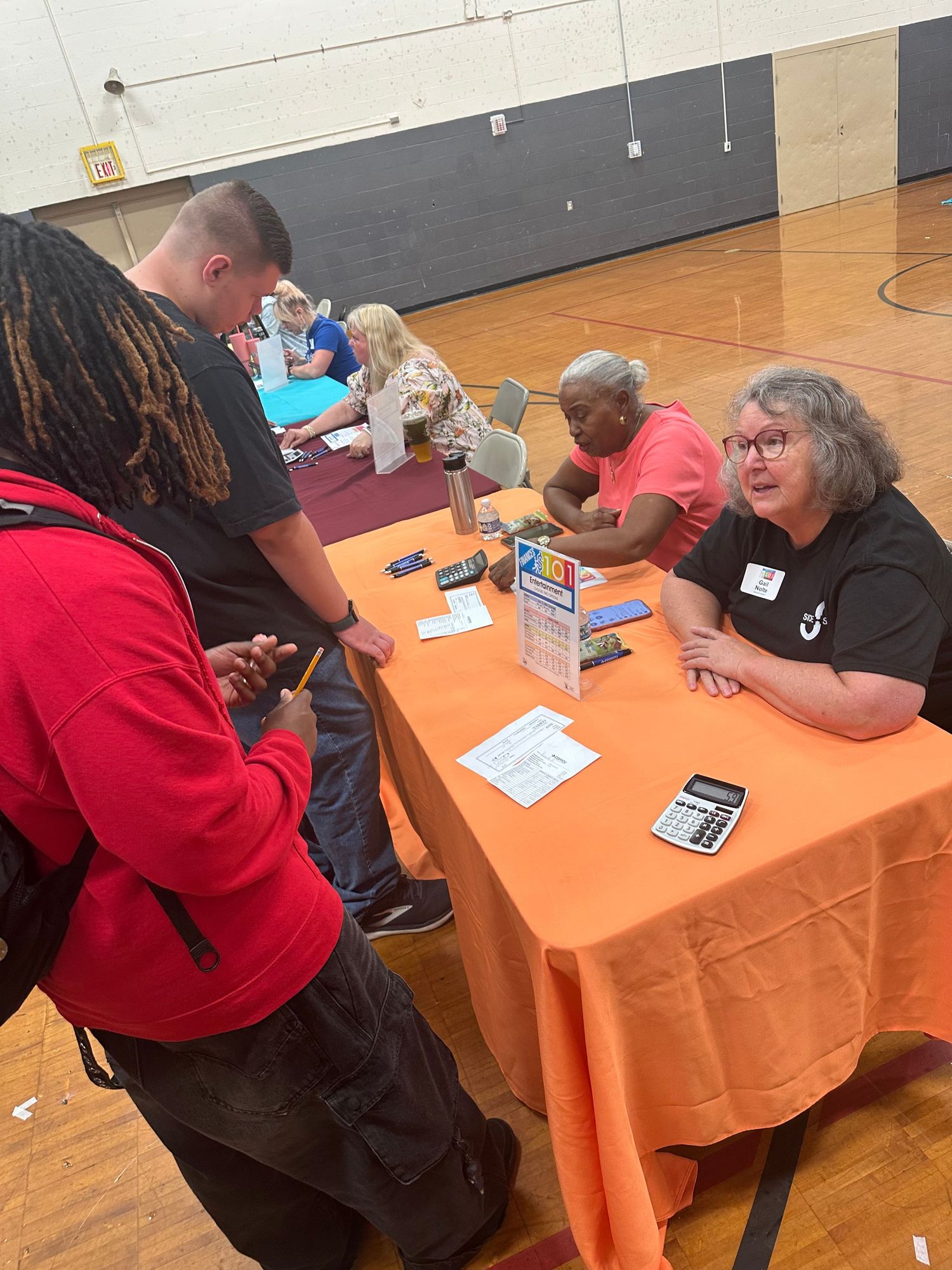 People gathered at orange-draped tables in a gymnasium for a community resource fair event.
