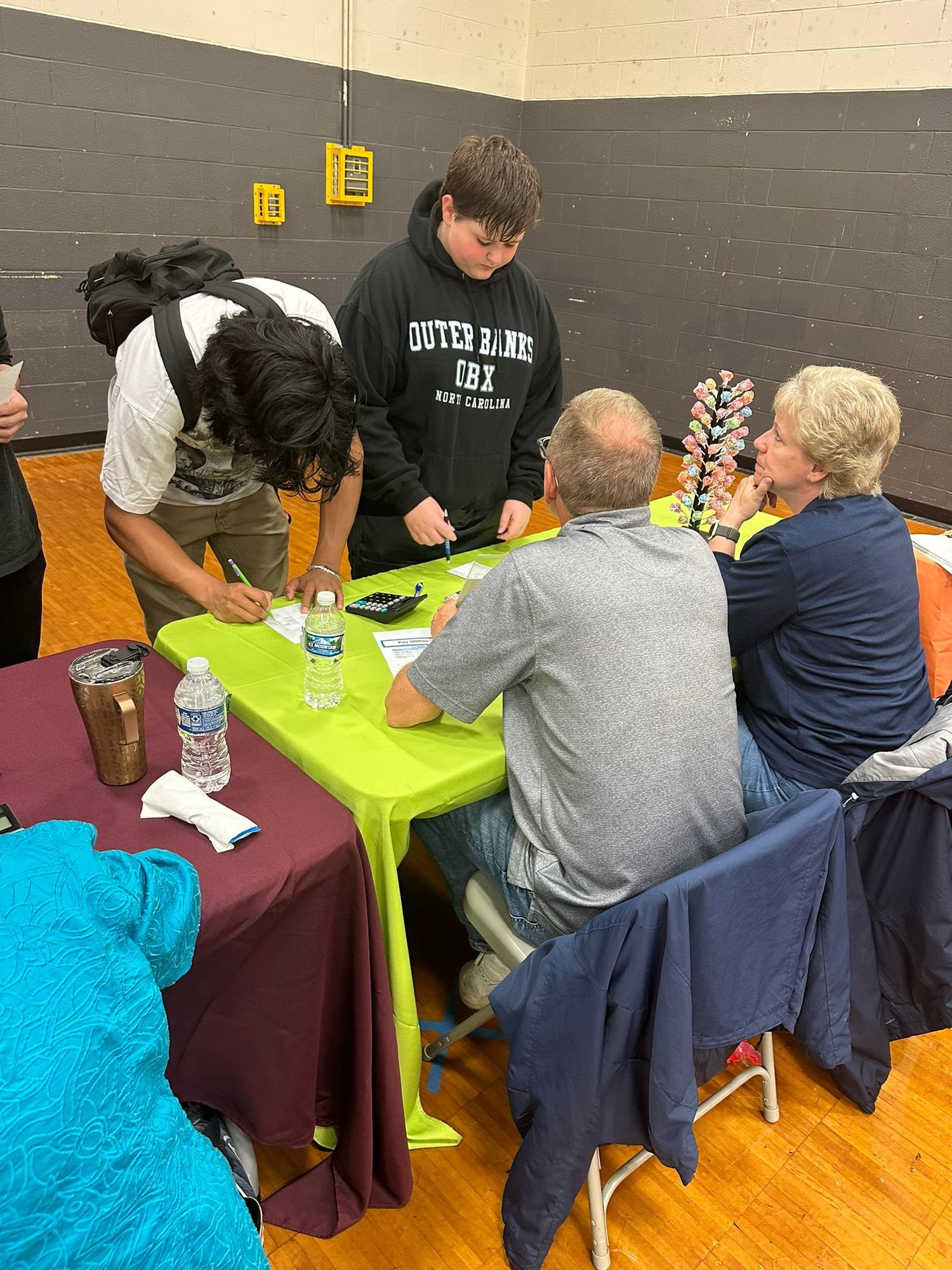 Students and adults collaborate around a bright green table in a school gymnasium during an educational activity.