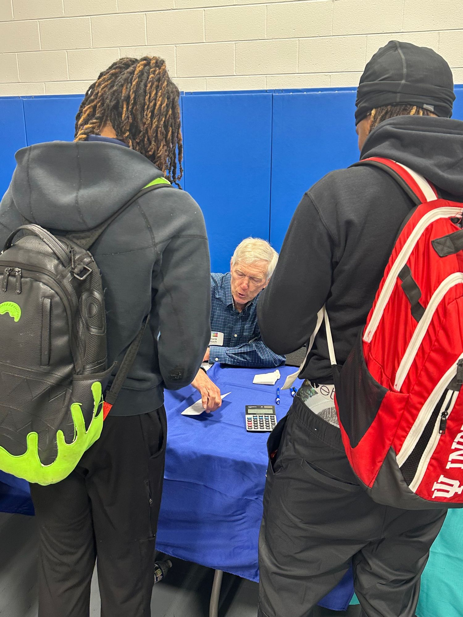 Two students with backpacks speak with an elderly man at a blue table during a school career or resource fair event.