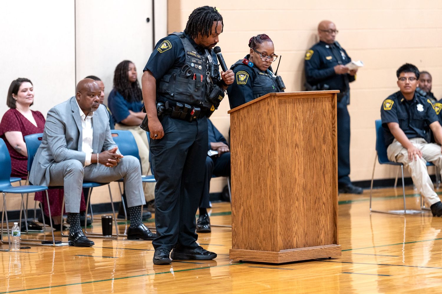 Two police officers speak at a podium during a community meeting in a gymnasium with attendees seated nearby.