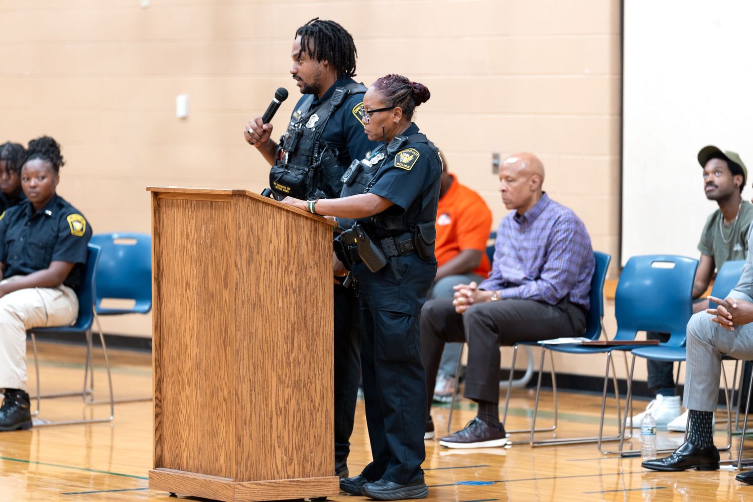Two police officers speak at a podium during a community meeting in a gymnasium with attendees seated in chairs.