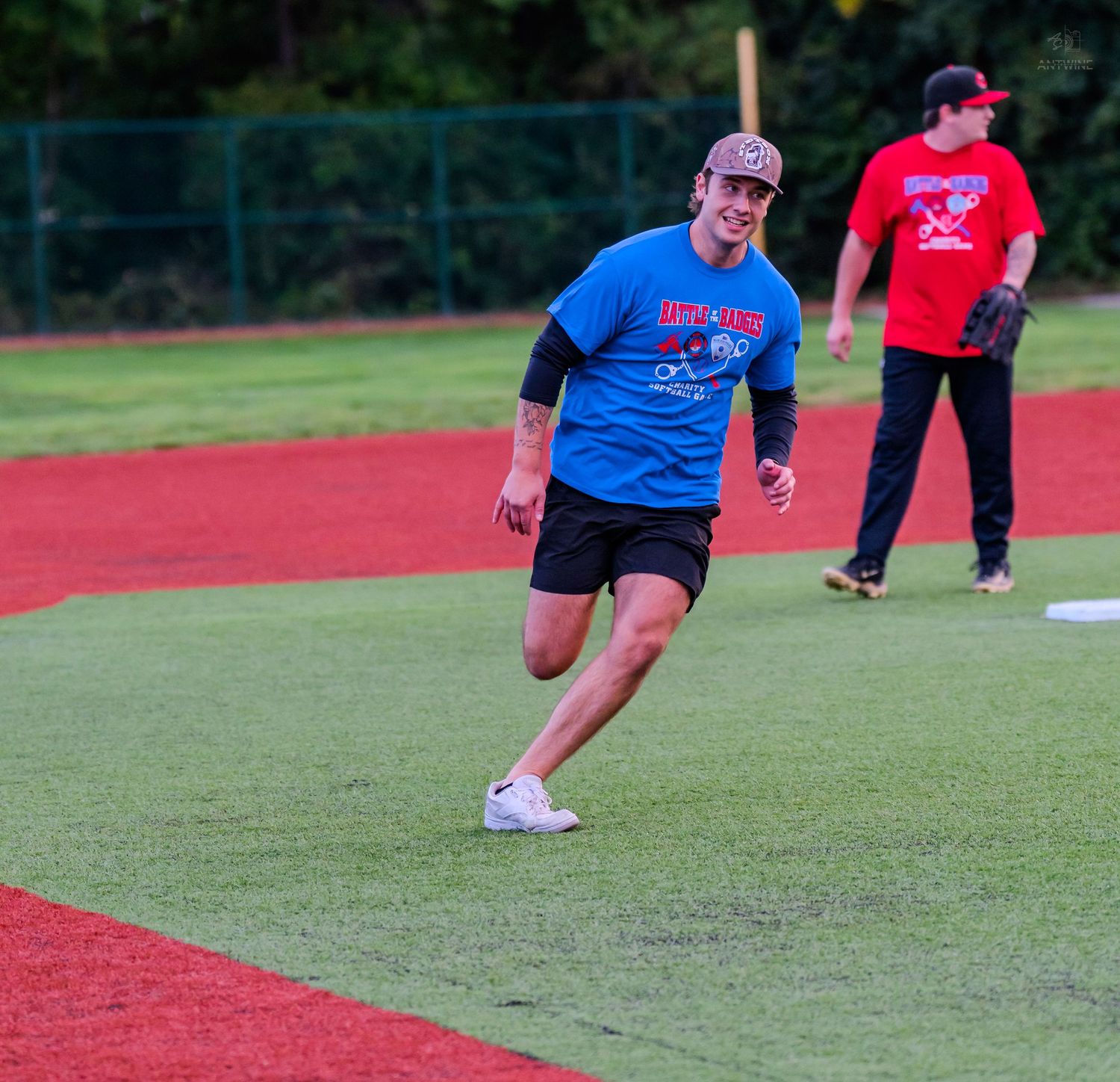 Smiling man in blue shirt running bases on baseball field with teammate in red uniform behind him.