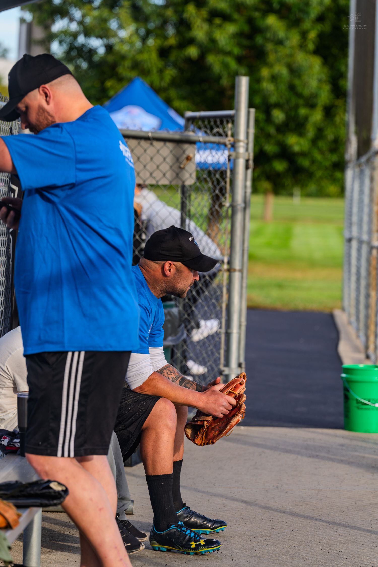 Two baseball players in blue shirts sit and stretch in a dugout during a softball game outdoors.