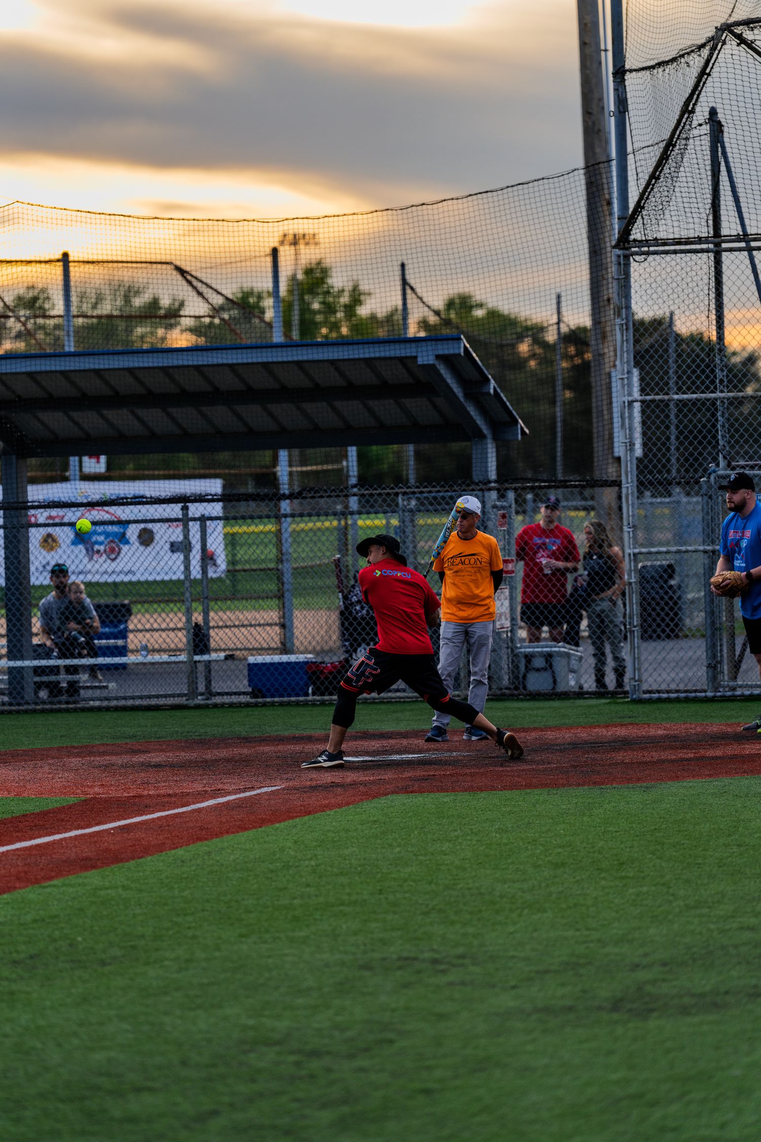 Softball batter in red jersey swinging at pitch during twilight game with orange sunset sky backdrop.