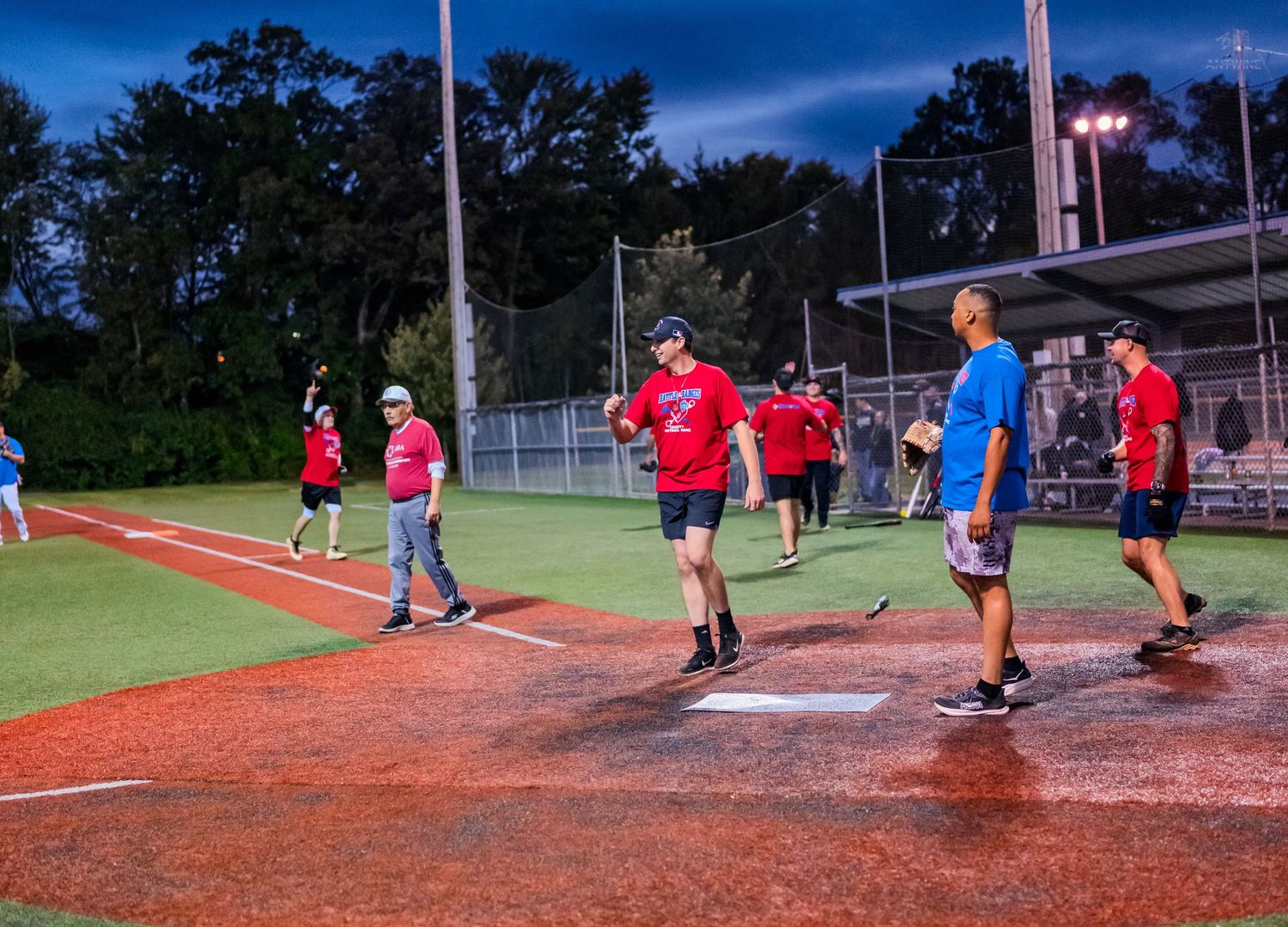 softball team in red shirts walks across illuminated red turf field during evening game.