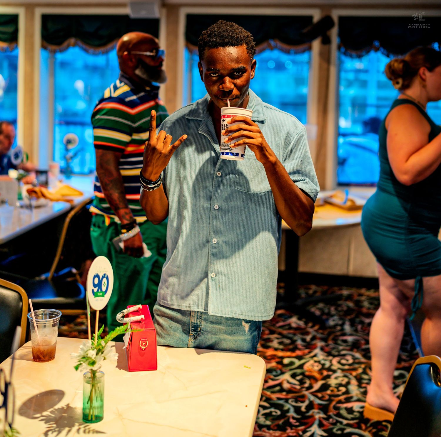 Young man sipping a drink at a table during an indoor riverboat party event.
