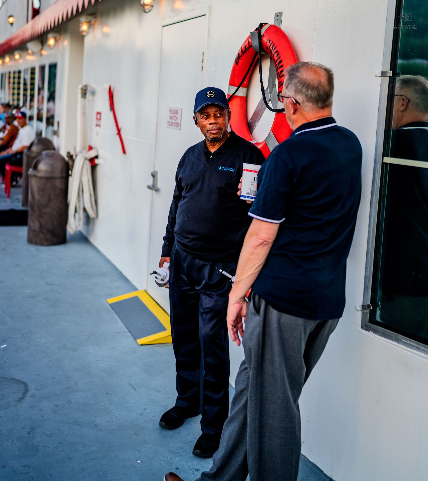 Two men in navy uniforms converse on a ship deck near a red life preserver ring.