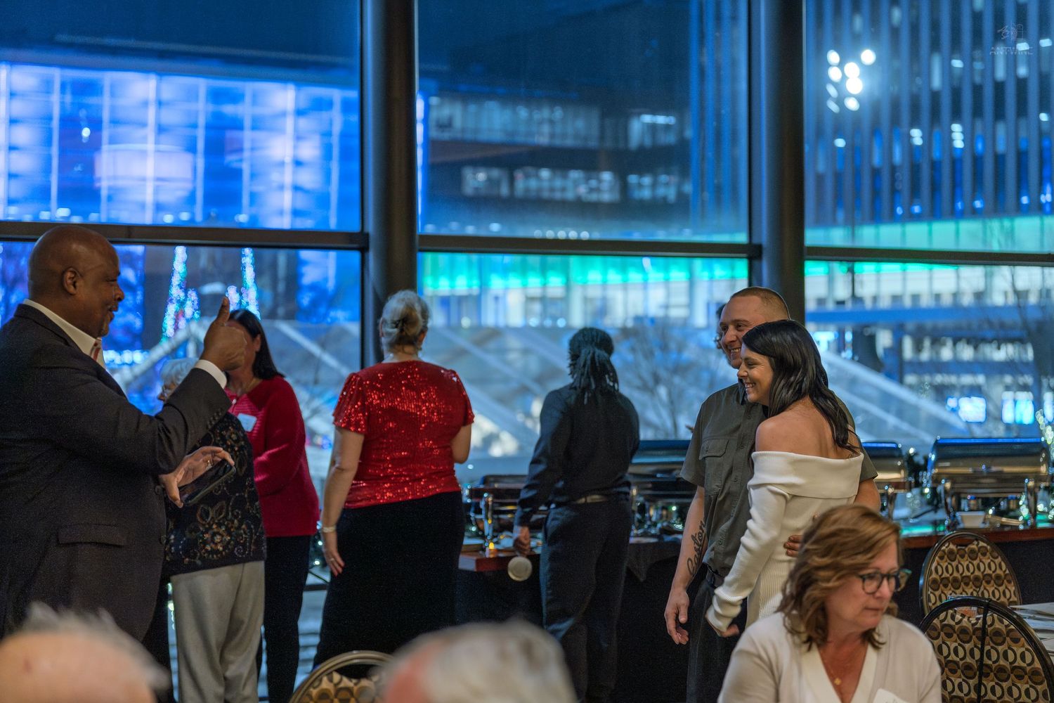 Elegantly dressed guests mingle at an upscale evening event with stunning city skyline views through floor-to-ceiling windows.