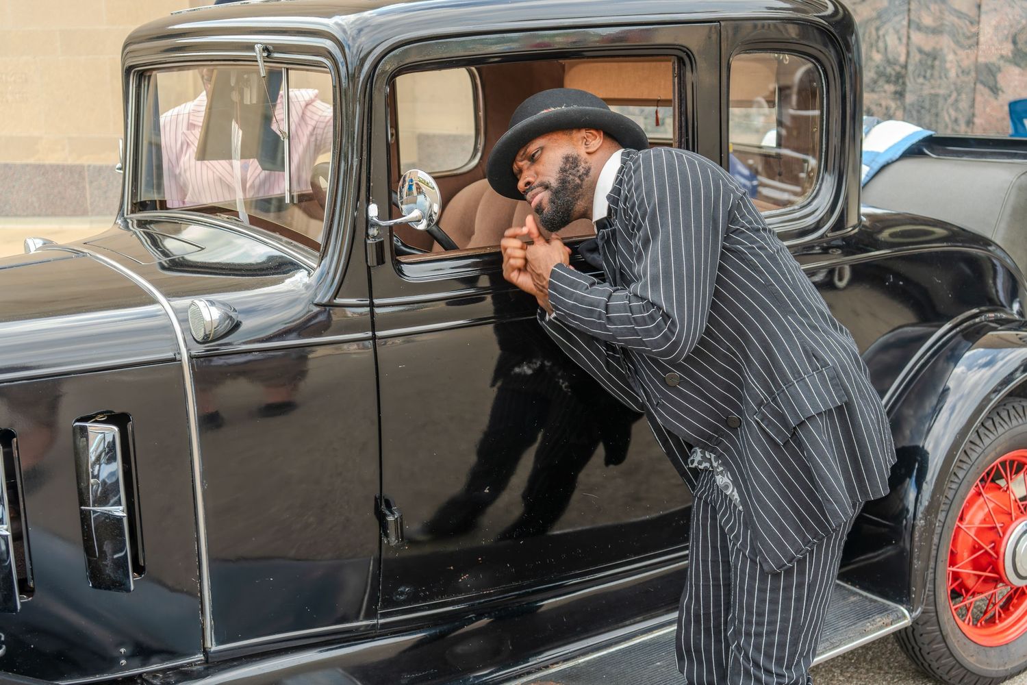 Man in pinstripe suit and beret examining a classic vintage black car at an outdoor automobile show.