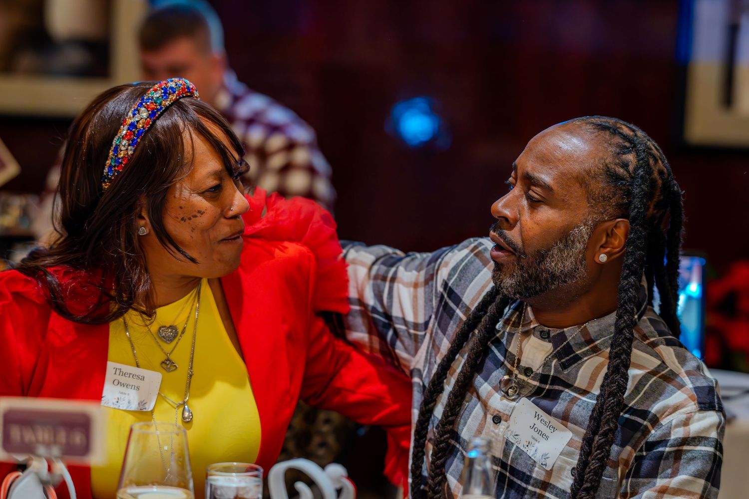 A man with dreadlocks in plaid shirt converses with a woman in red jacket at an indoor event gathering.