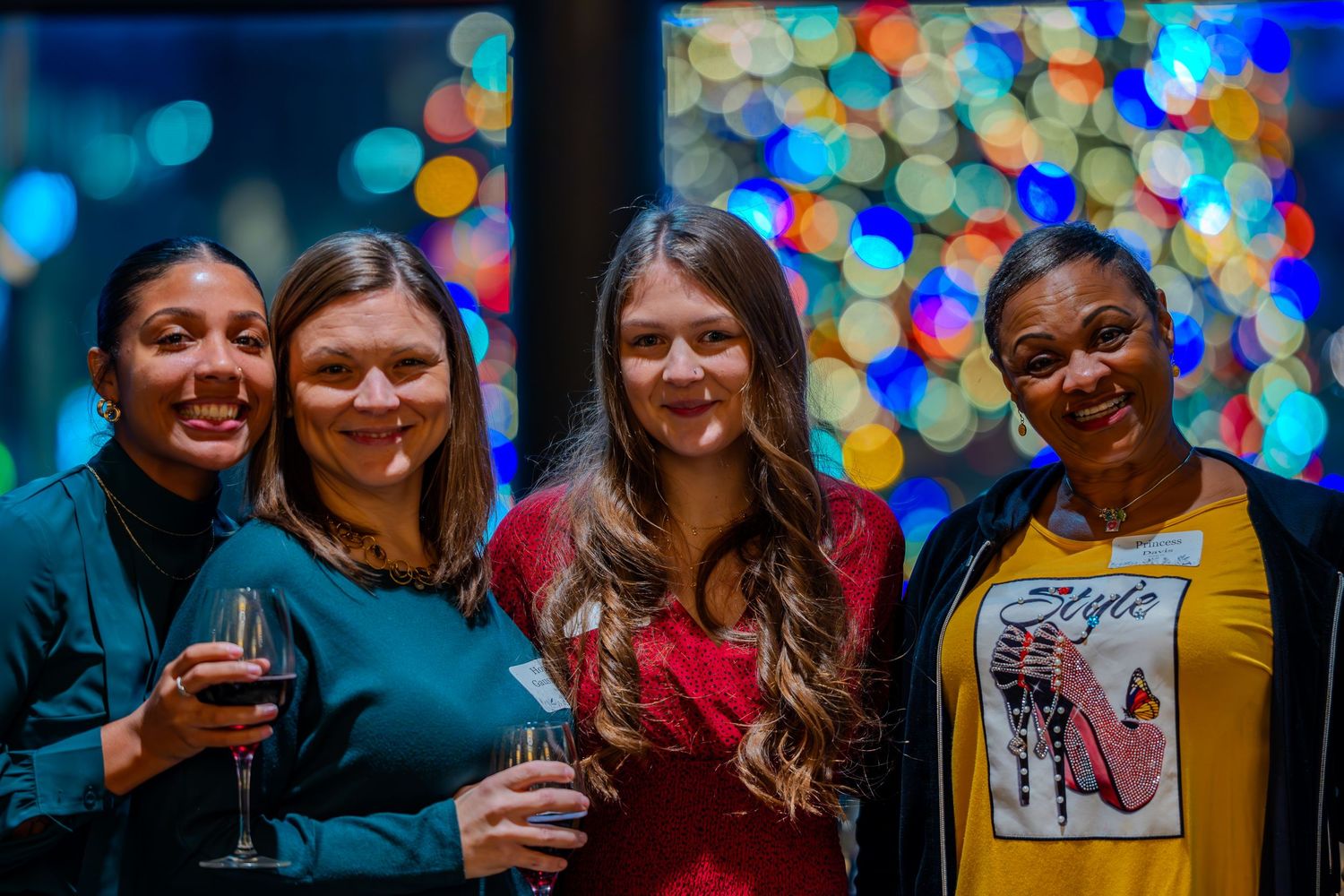 Four women smiling together at a festive holiday party with colorful bokeh lights in the background.