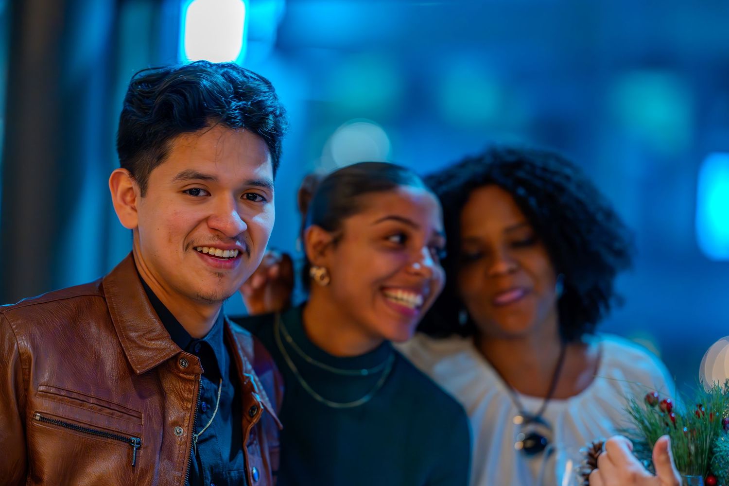 Three smiling friends posing together at a vibrant nighttime event, wearing casual jackets in blue-lit surroundings.