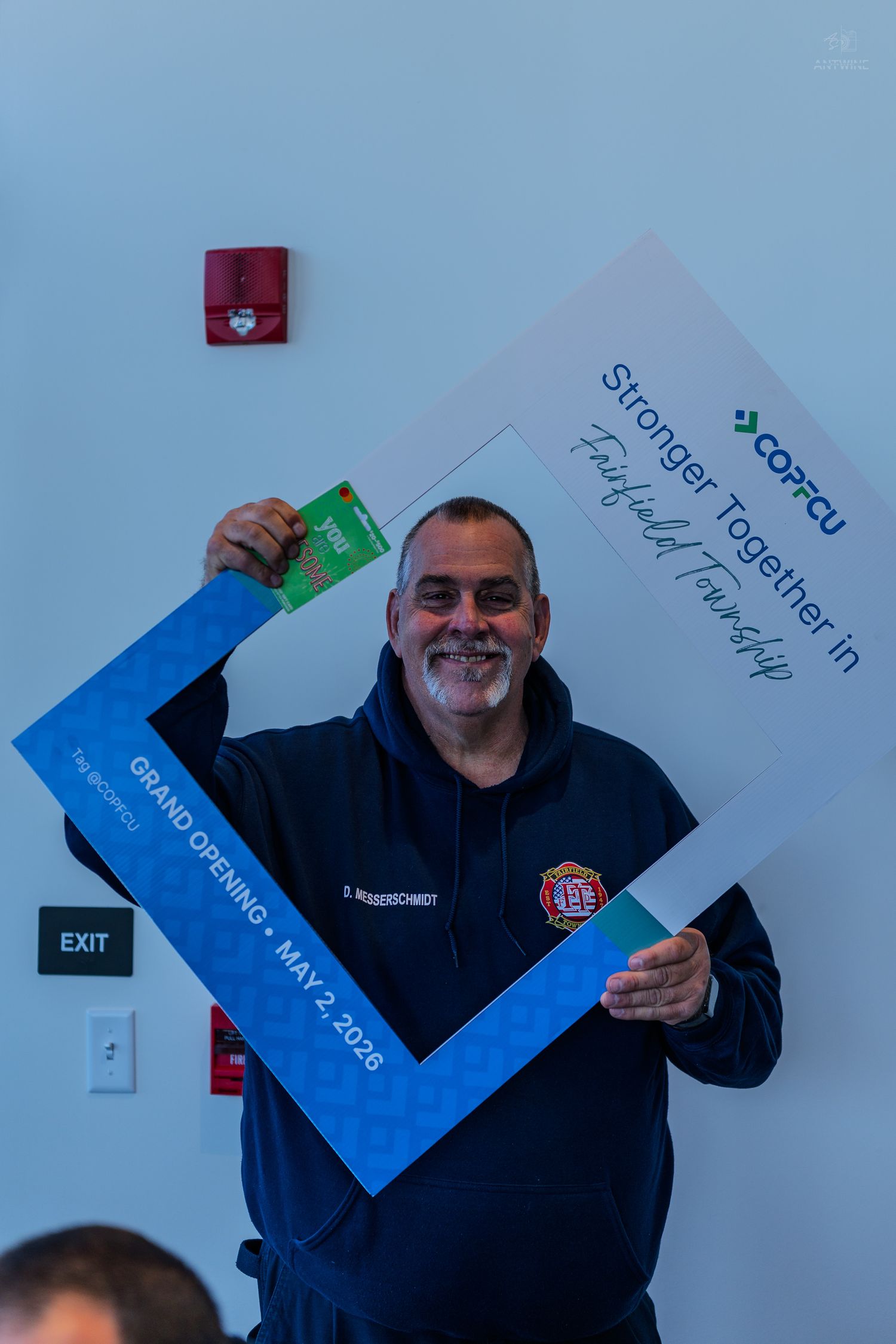 Man in firefighter hoodie holding COPFCU Stronger Together grand opening frame sign on May 2, 2026.