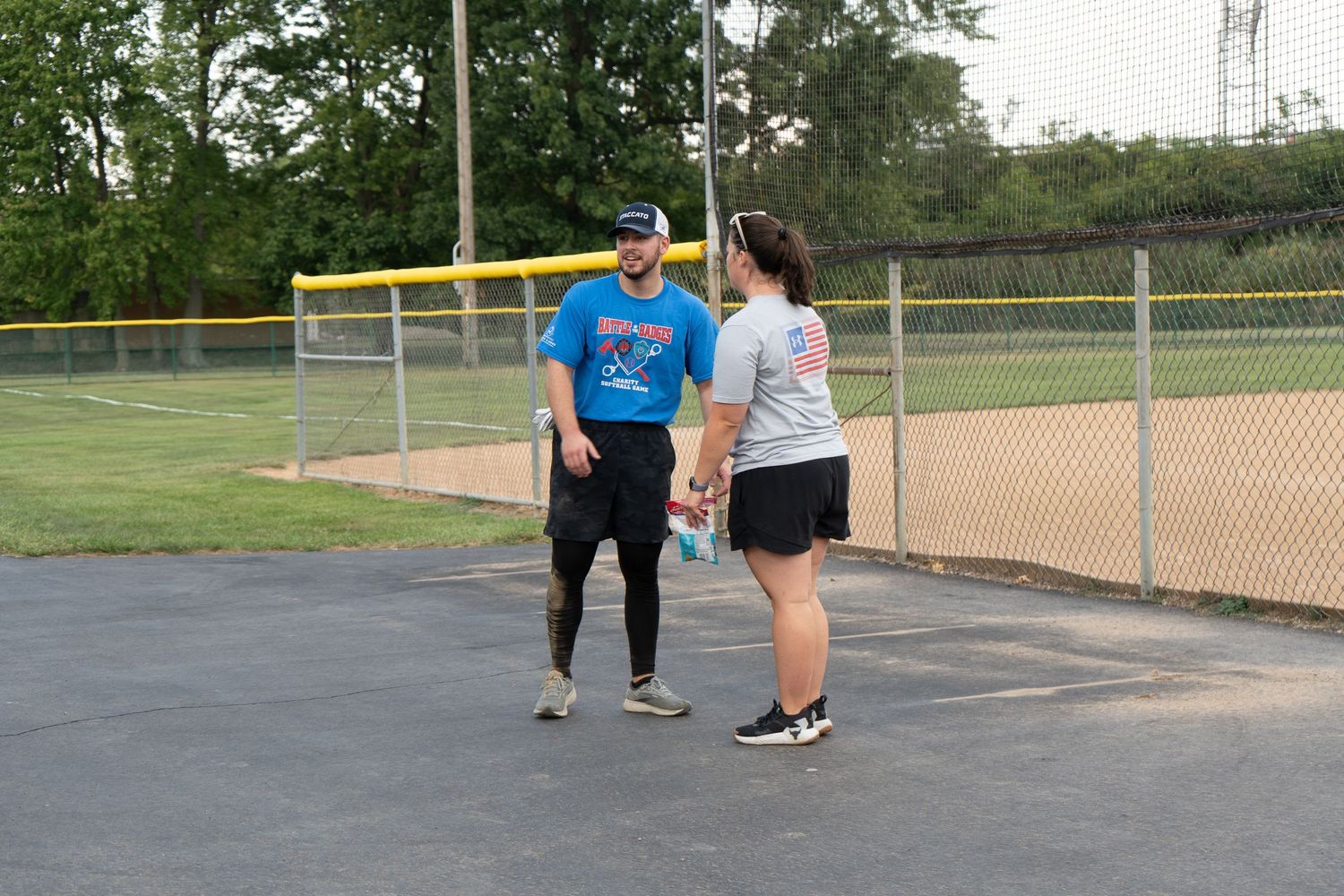 A man and woman holding hands and talking near a chain-link fence at an outdoor baseball field.