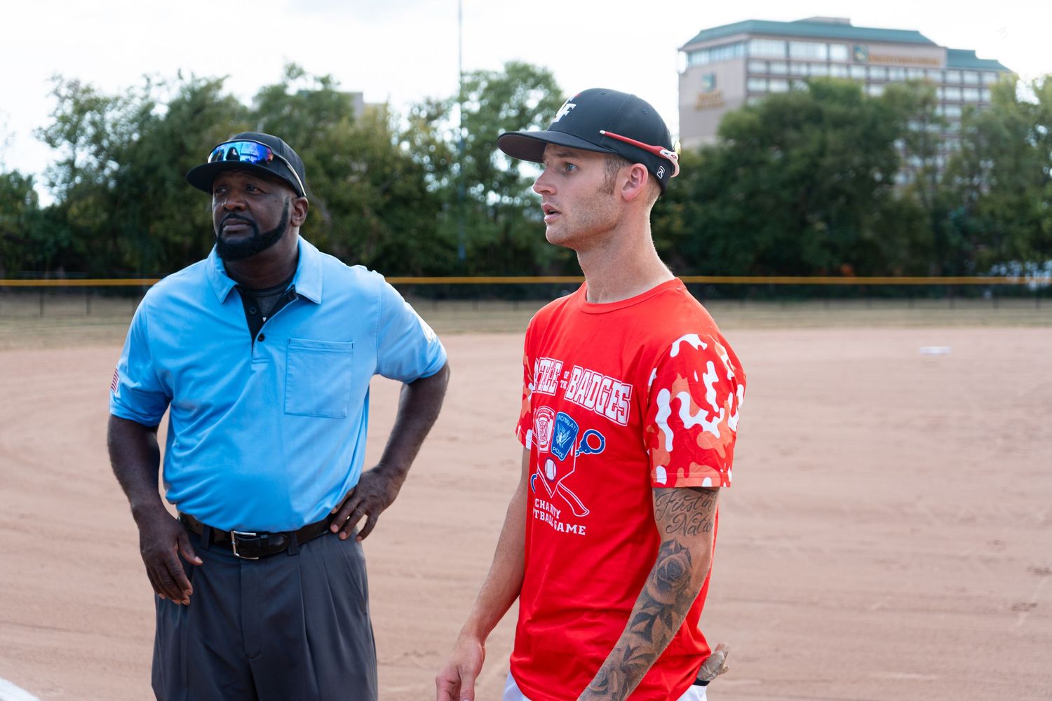 A softball player in a red jersey stands beside an umpire in a blue shirt on the field.