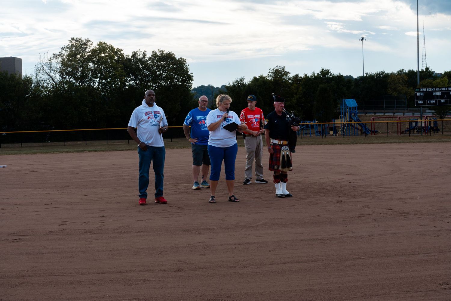 Four people stand on a dirt softball field at dusk near a bagpiper during an outdoor ceremony.