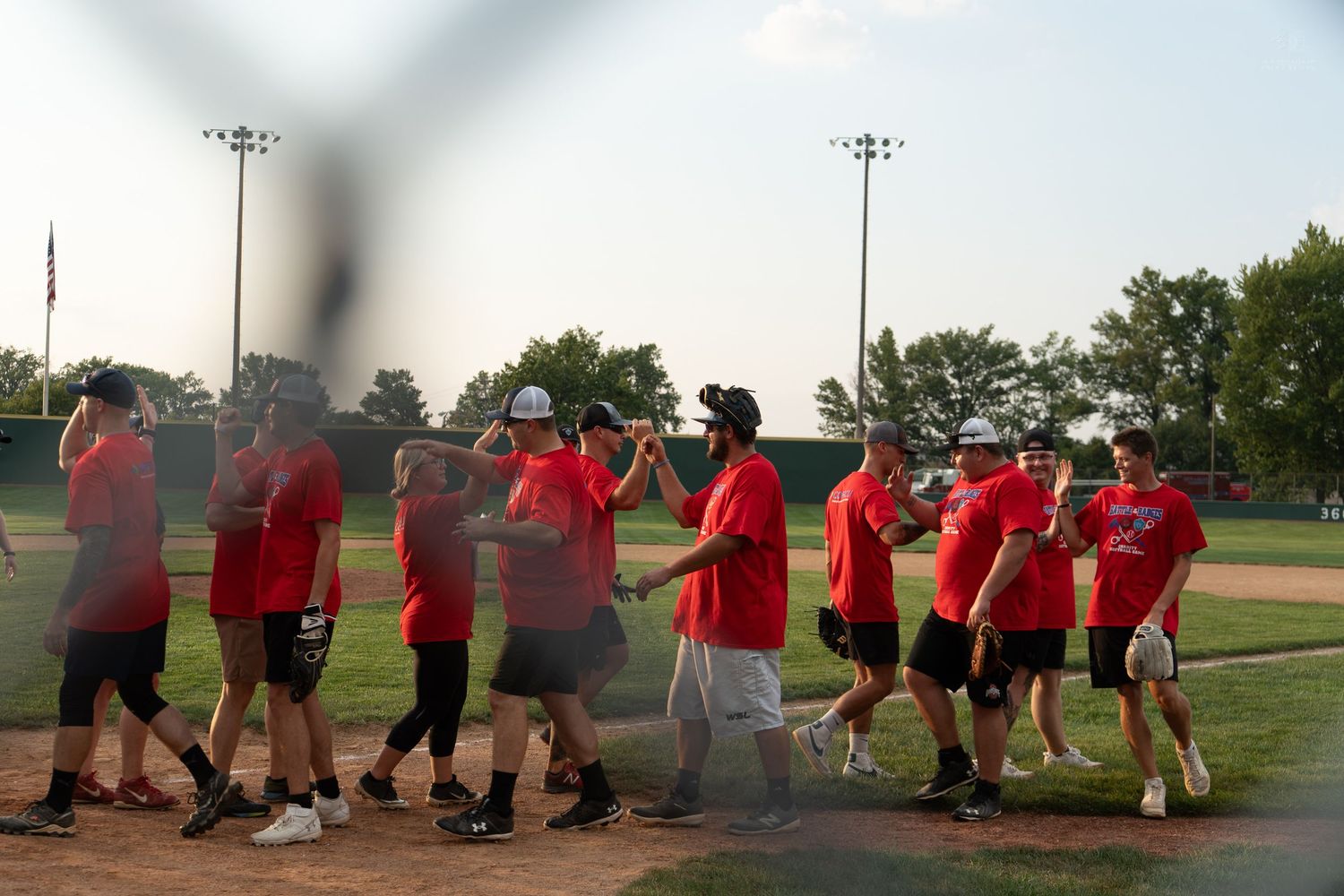 Baseball team in red shirts celebrating on field at dusk, with stadium lights and trees in background.