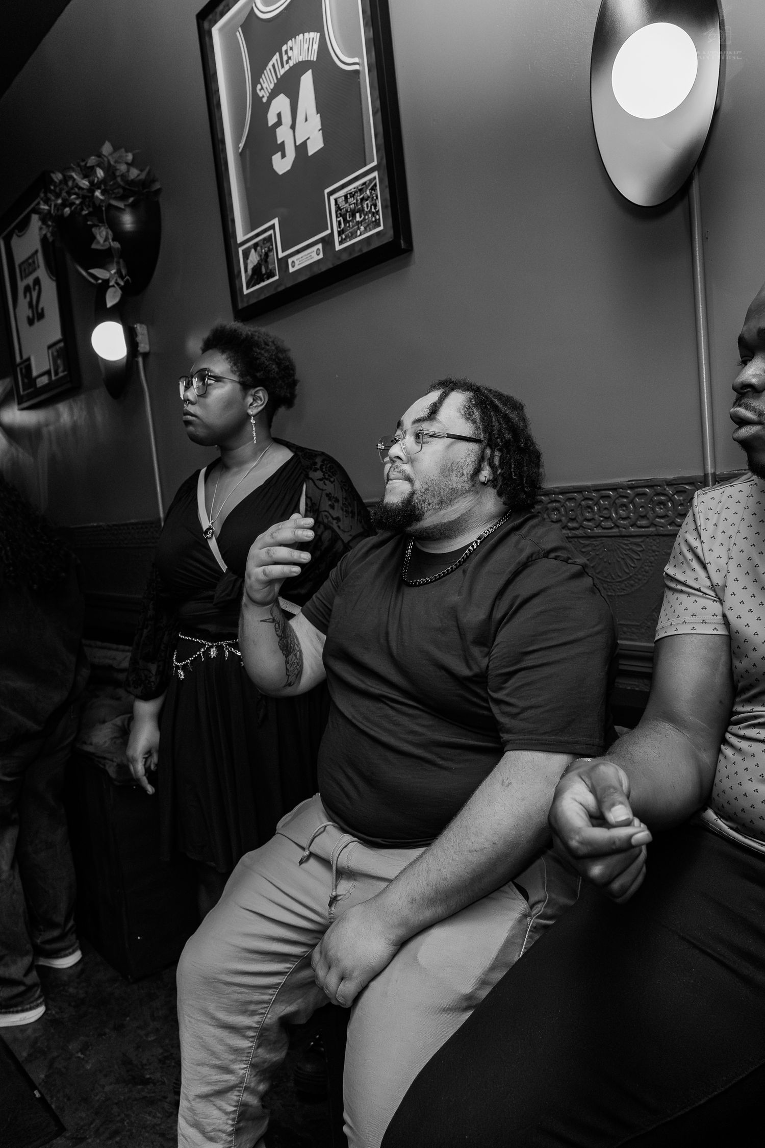 Black and white photo of people gathered at a bar, man in t-shirt holding drink with framed jersey on wall.