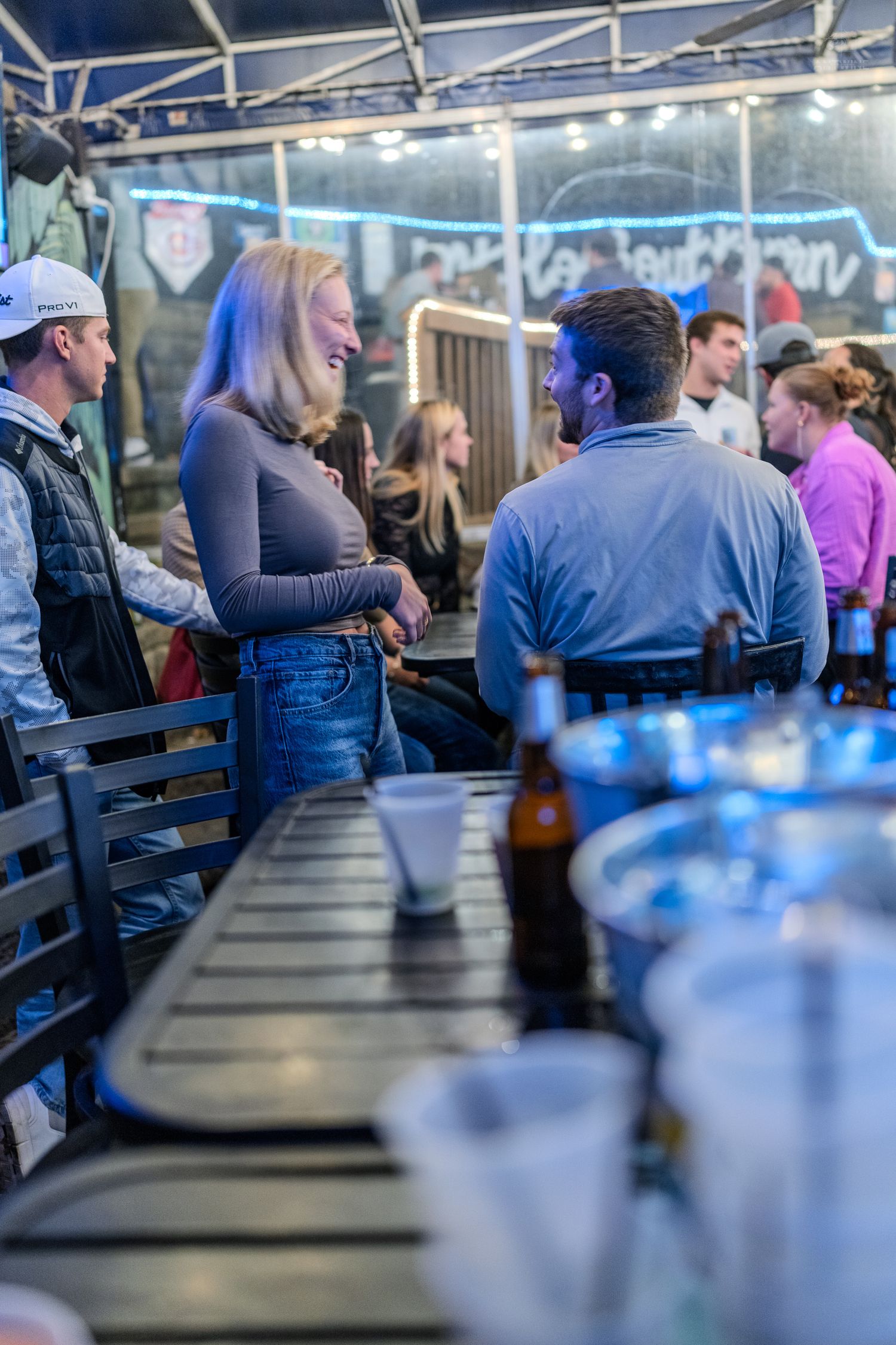 People socializing at an outdoor bar patio at night with string lights, beer bottles, and cups on a wooden table.