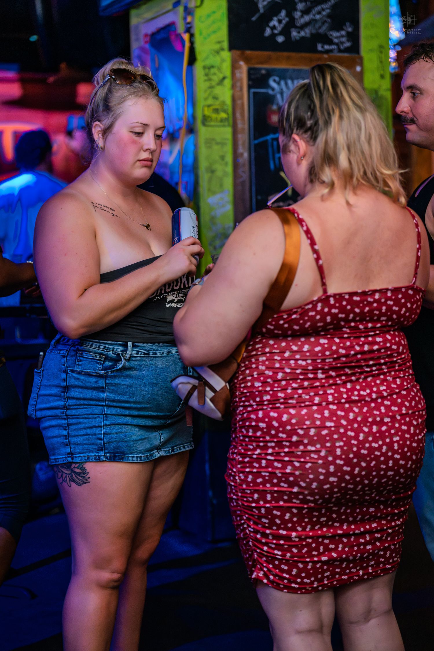 Two women socializing in a colorful bar, one in denim shorts and one in a red dress.