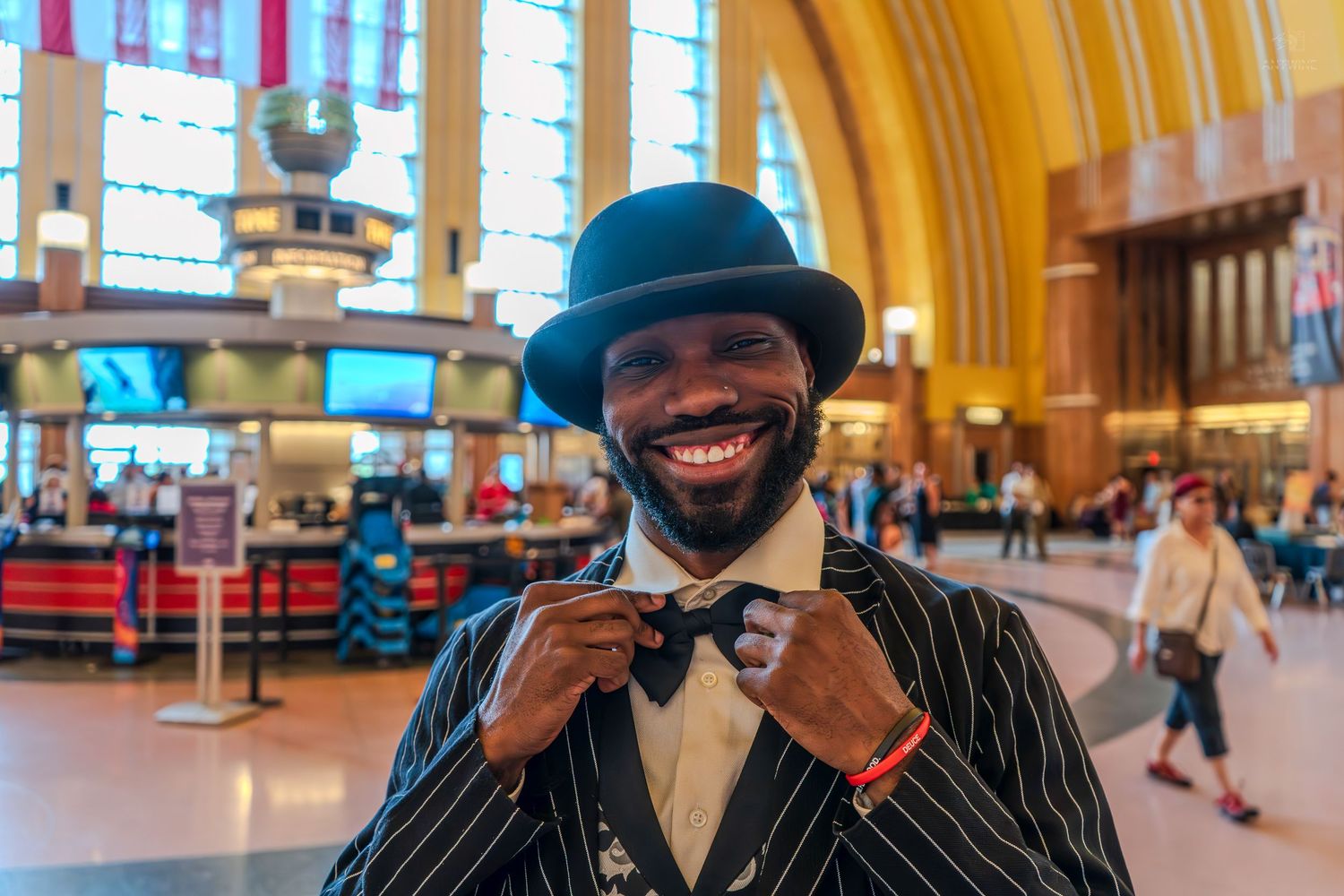 Smiling man in pinstripe suit and bowler hat adjusting bow tie inside a grand train station.