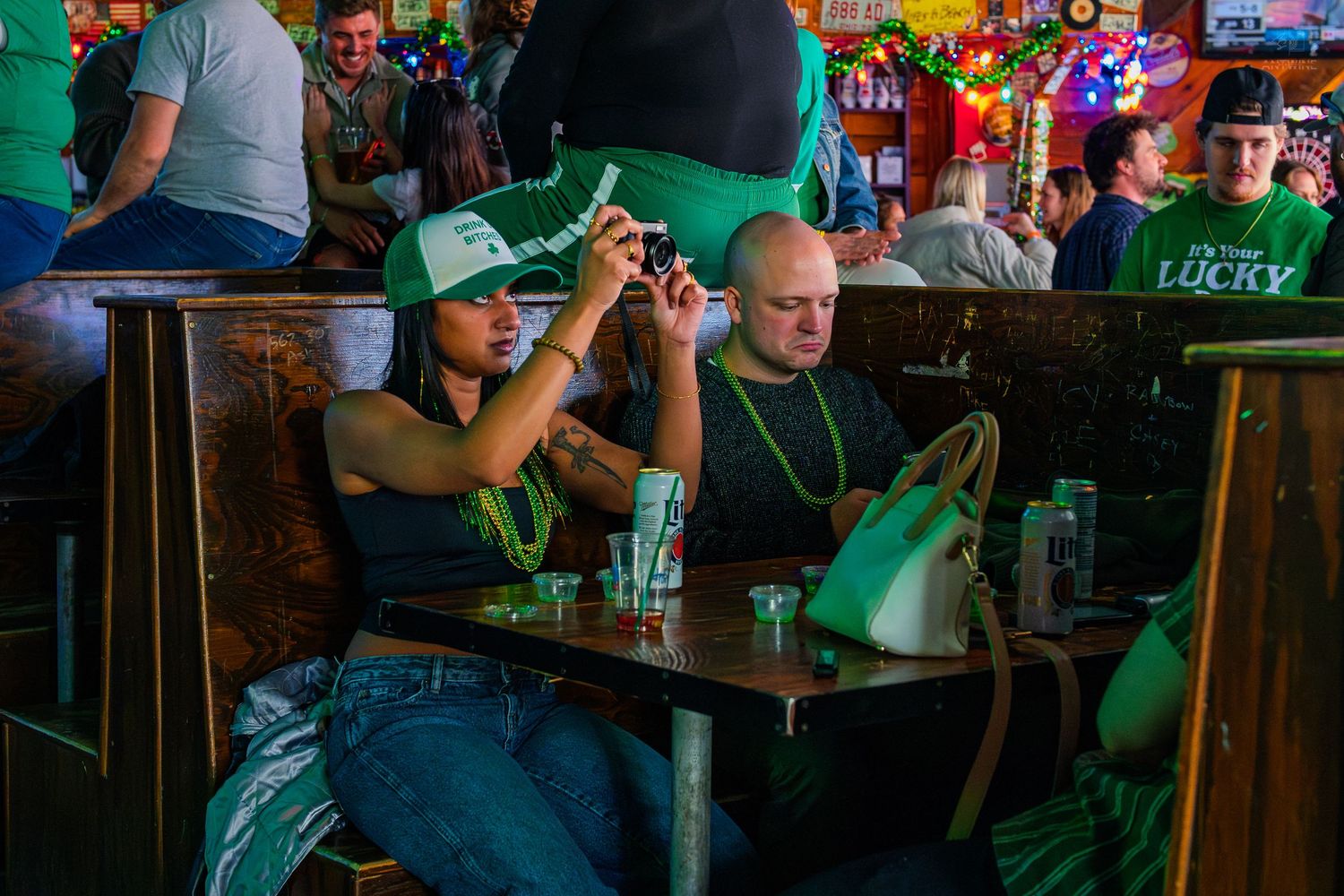 People celebrating St. Patrick's Day at a bar, wearing green outfits, with a woman taking a photo on her phone.
