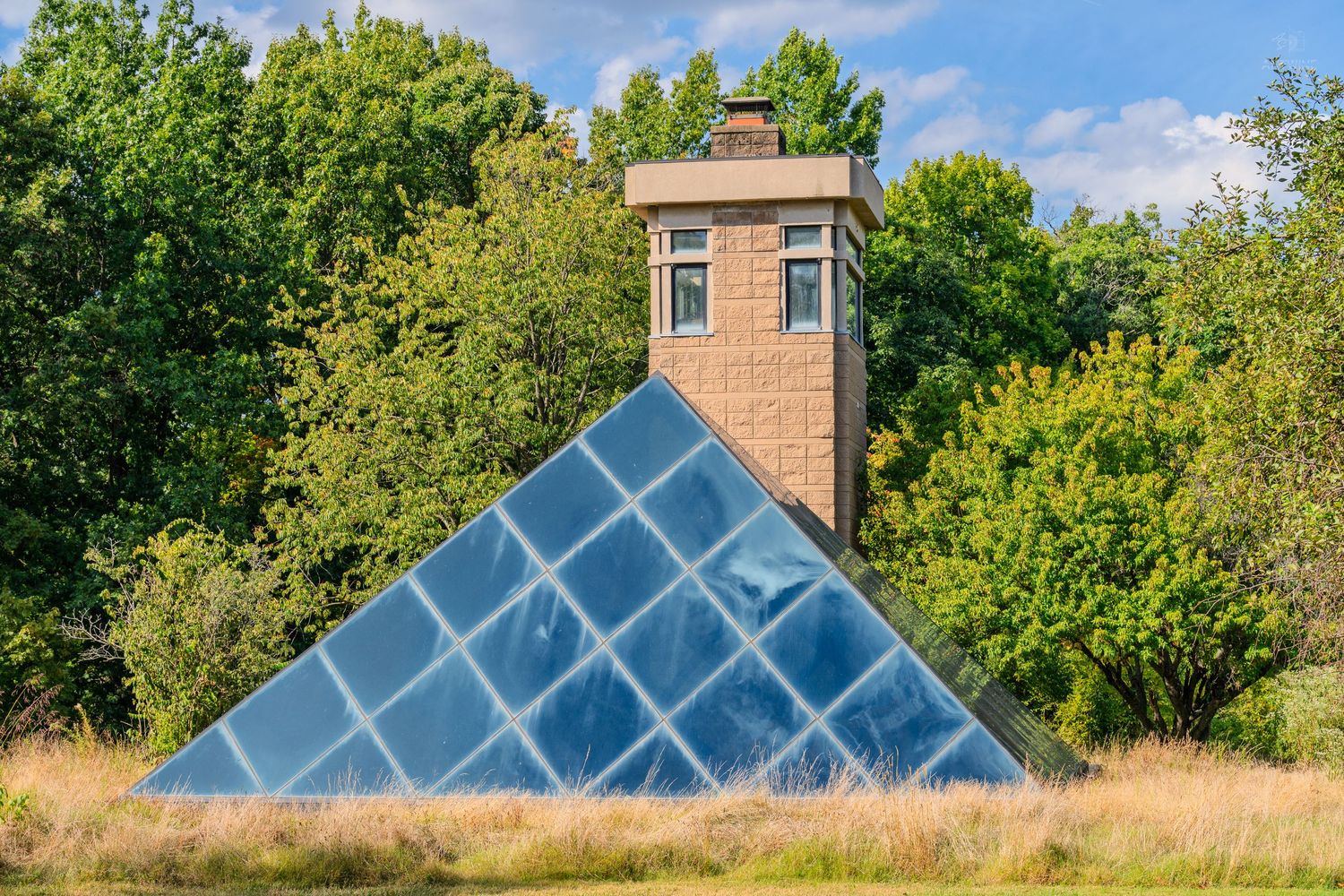 Glass pyramid structure beside a brick tower surrounded by lush green trees in an open field.