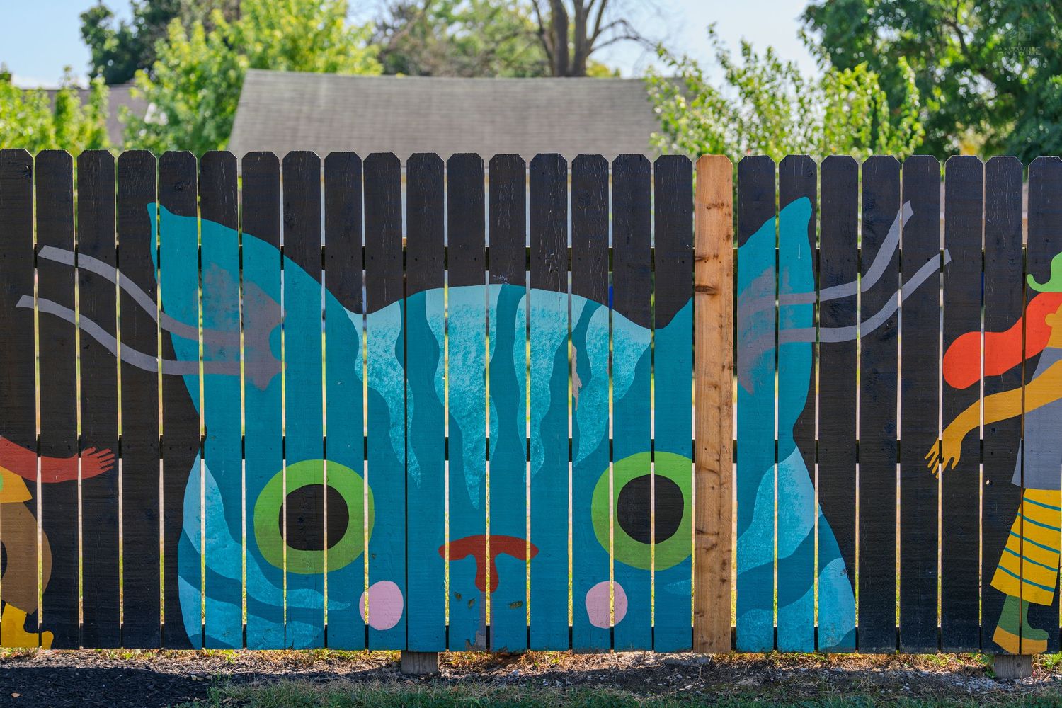 Colorful mural of a teal cartoon cat painted on a wooden backyard fence in bright sunlight.