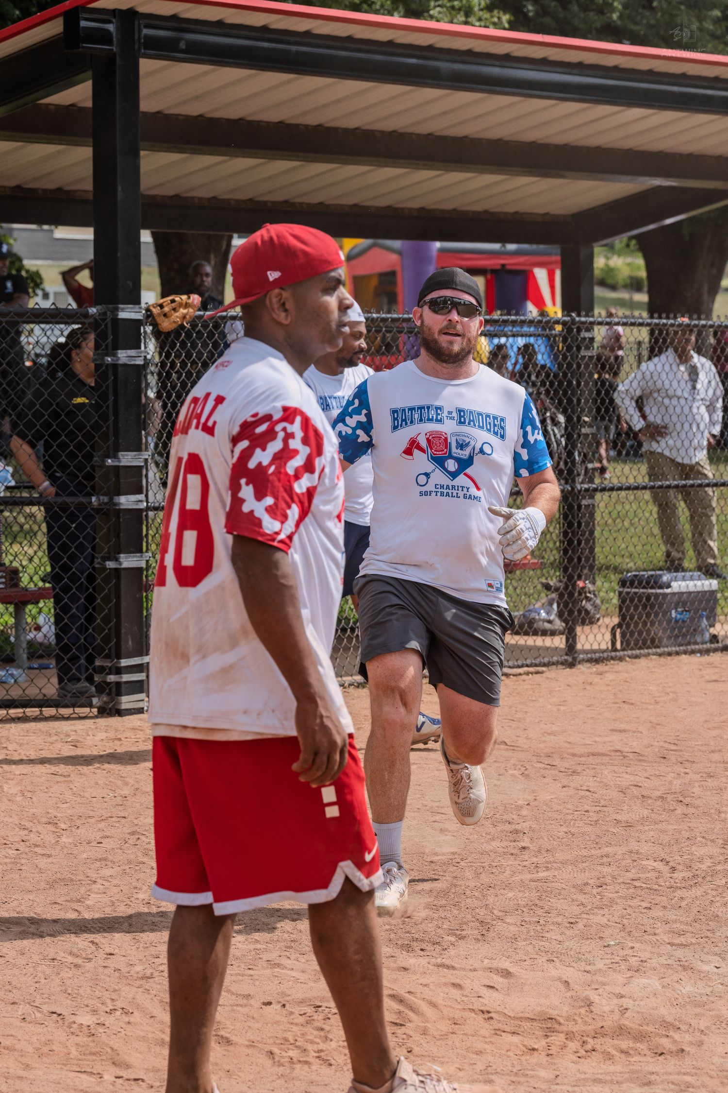 Two men in softball uniforms on a dirt field during a Battle of the Badges charity softball game outdoors.