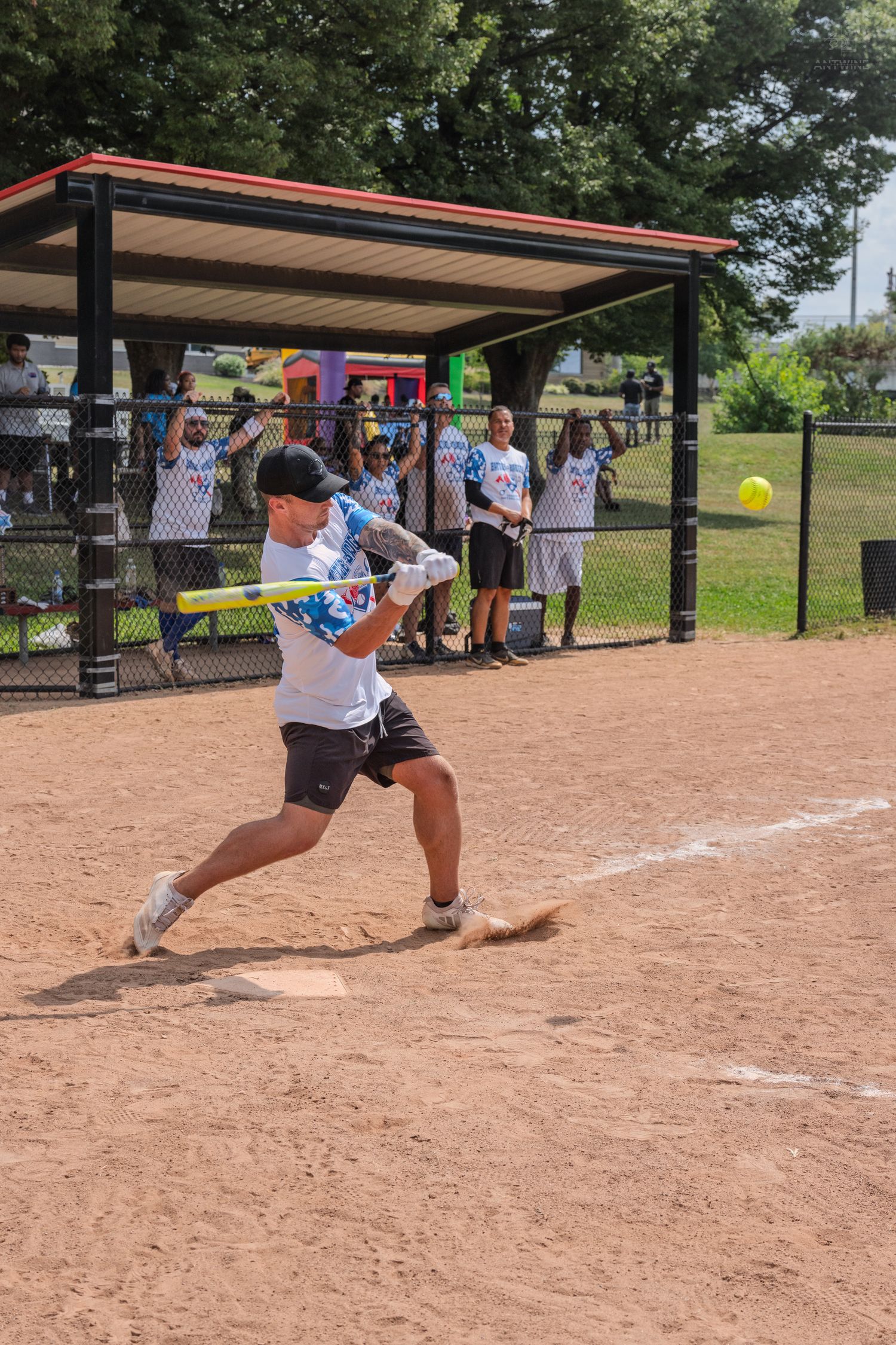 Male softball player swings bat at yellow ball during charity softball game near dugout.