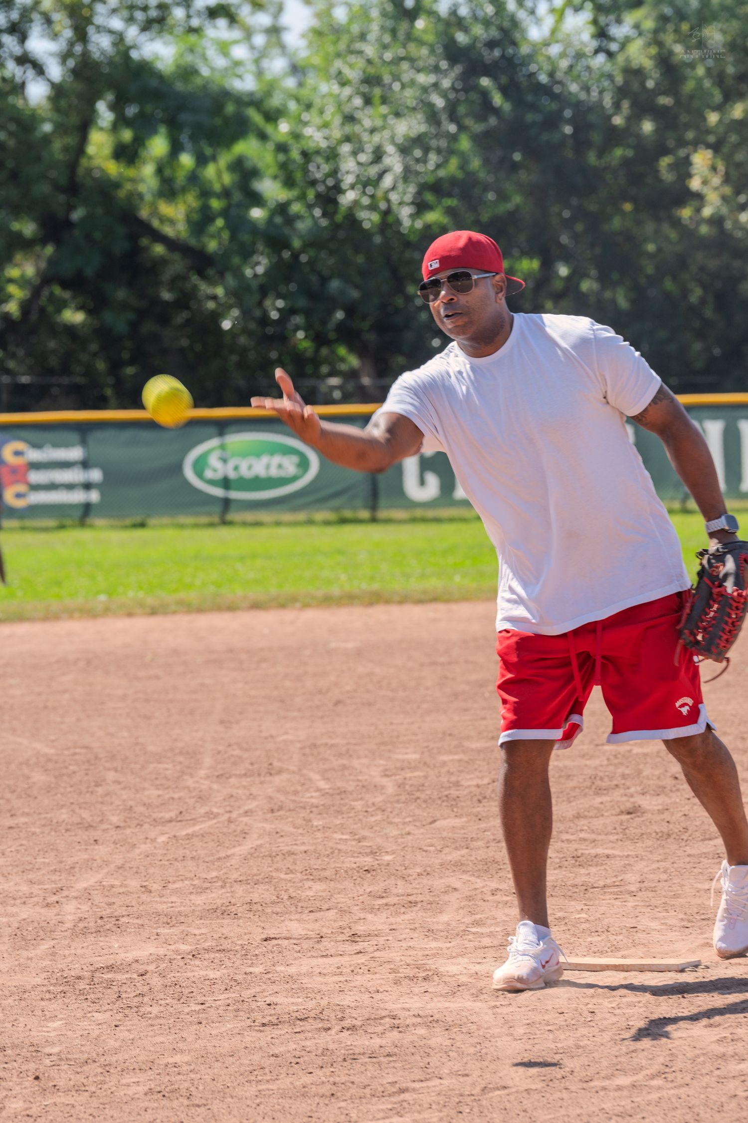 Man in red cap and white shirt throwing a yellow softball on a dirt infield during an outdoor recreational game.