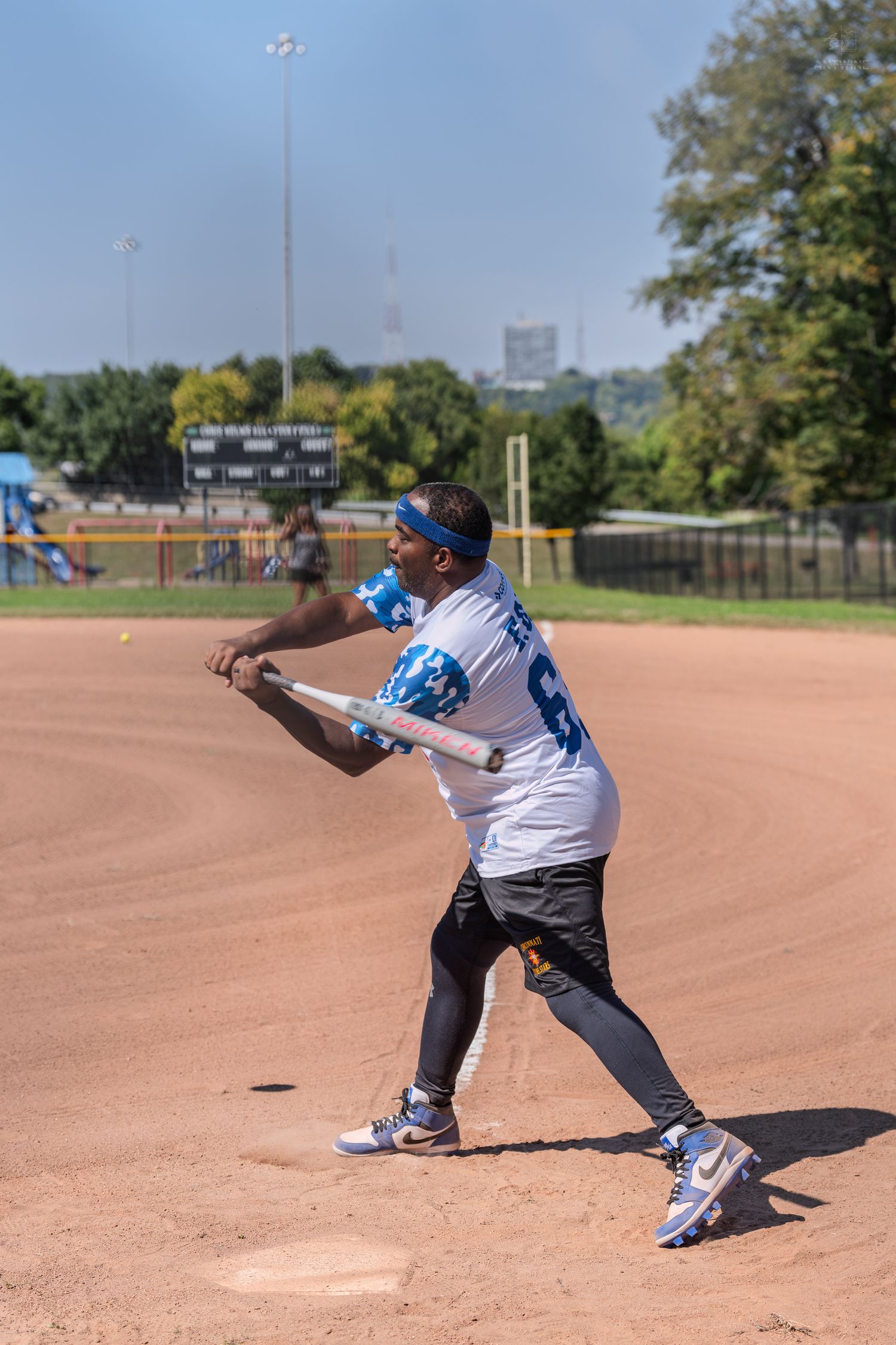 A man in a white jersey swings a baseball bat during a softball game on a sunny day at an outdoor diamond.