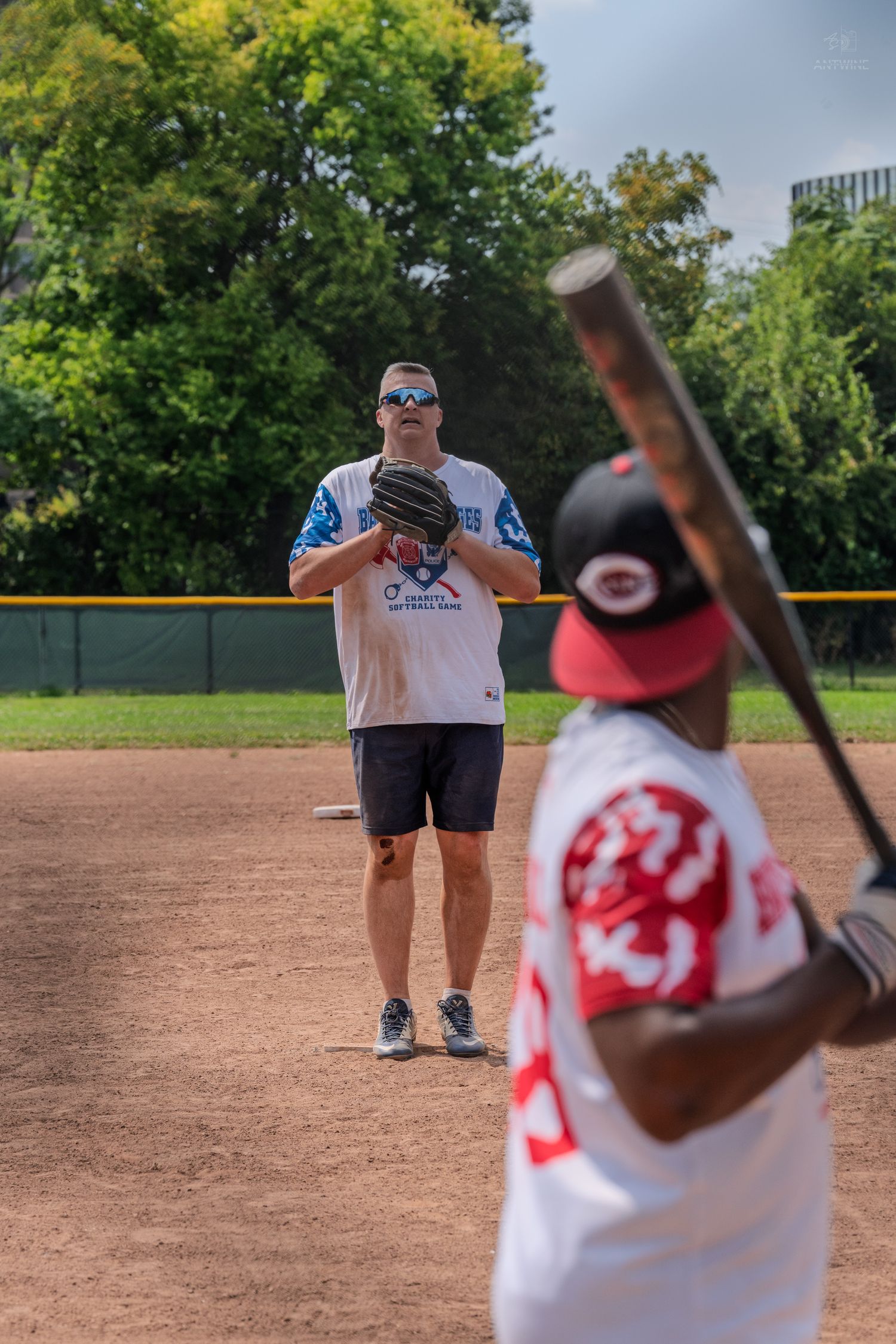 Baseball pitcher in blue jersey faces batter holding bat on outdoor dirt field with green trees.
