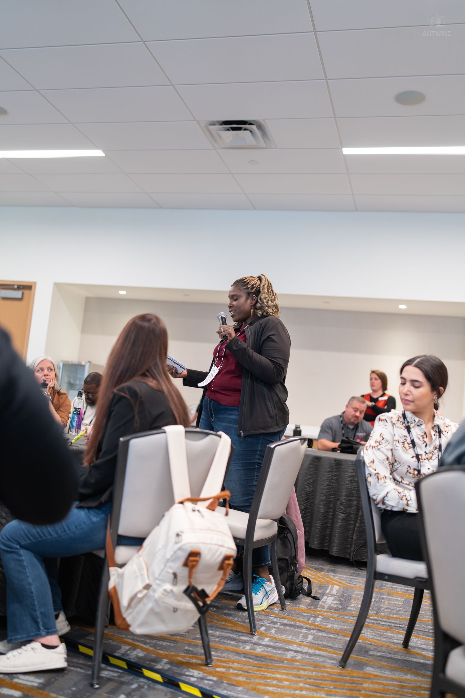 Woman speaking into a microphone while standing in a conference room during a professional event.