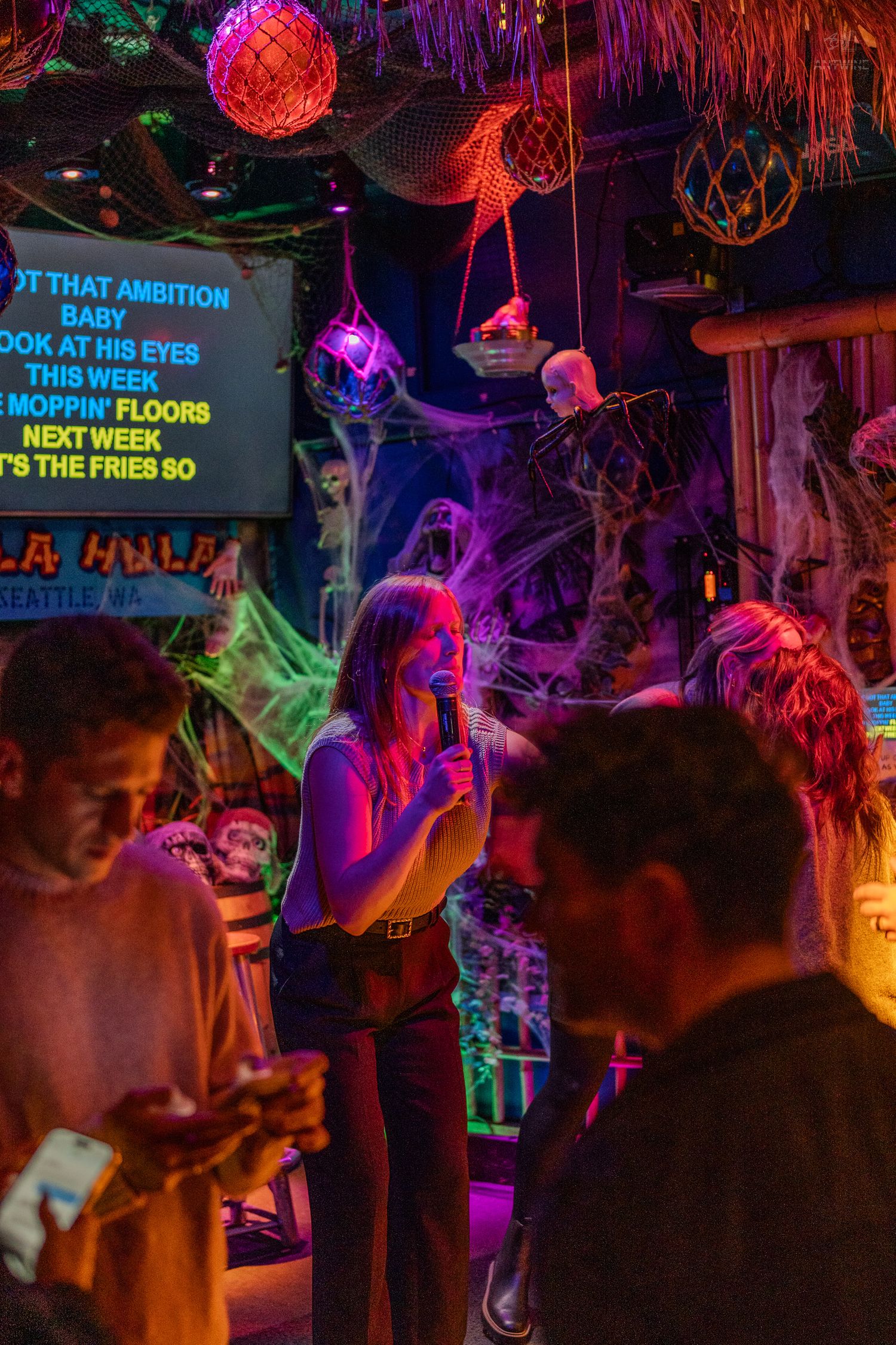 Woman singing karaoke on a neon-lit Halloween-decorated stage at a tropical-themed bar.
