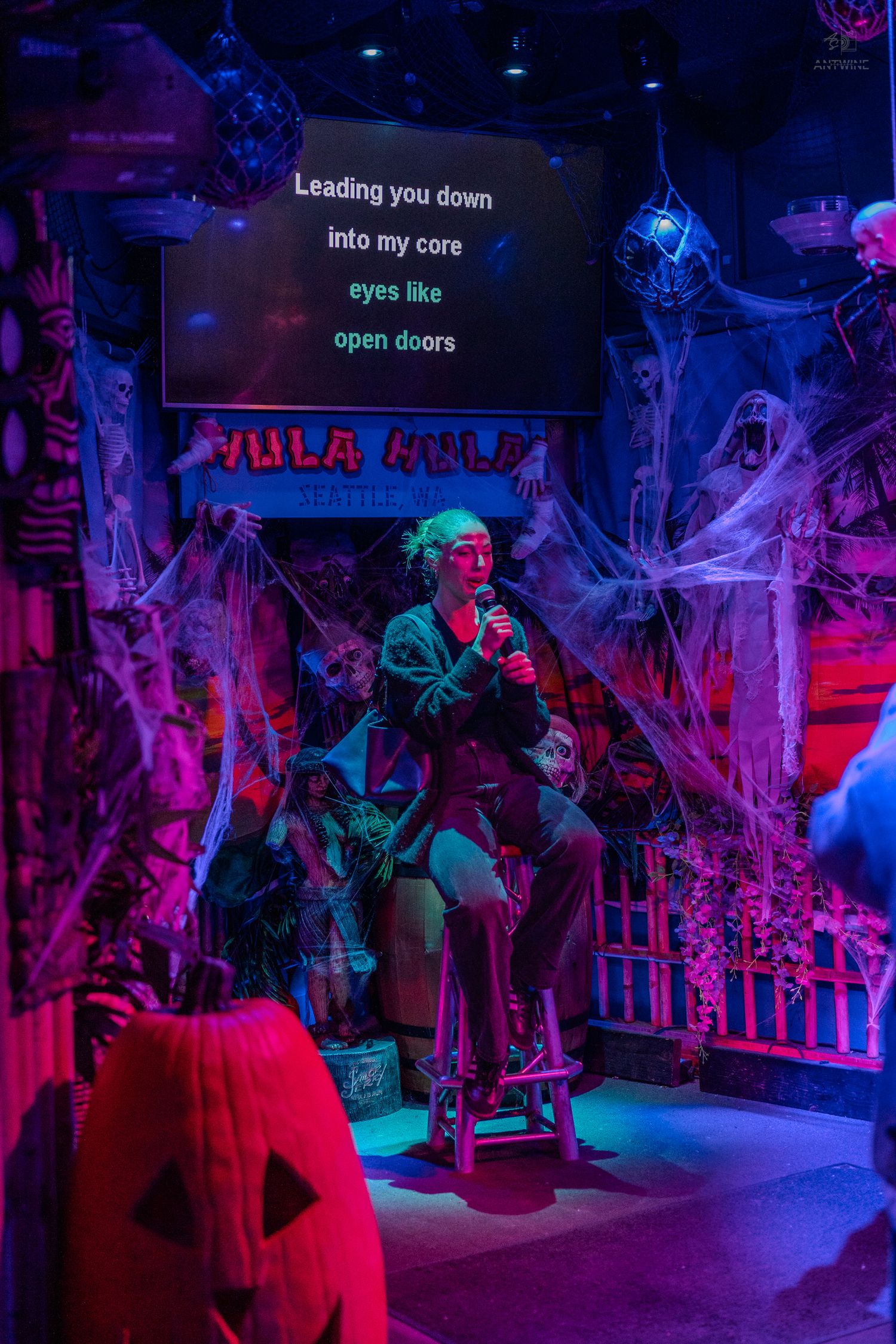 Singer performing karaoke seated on stool on spooky Halloween-decorated stage at Hula Hula bar.